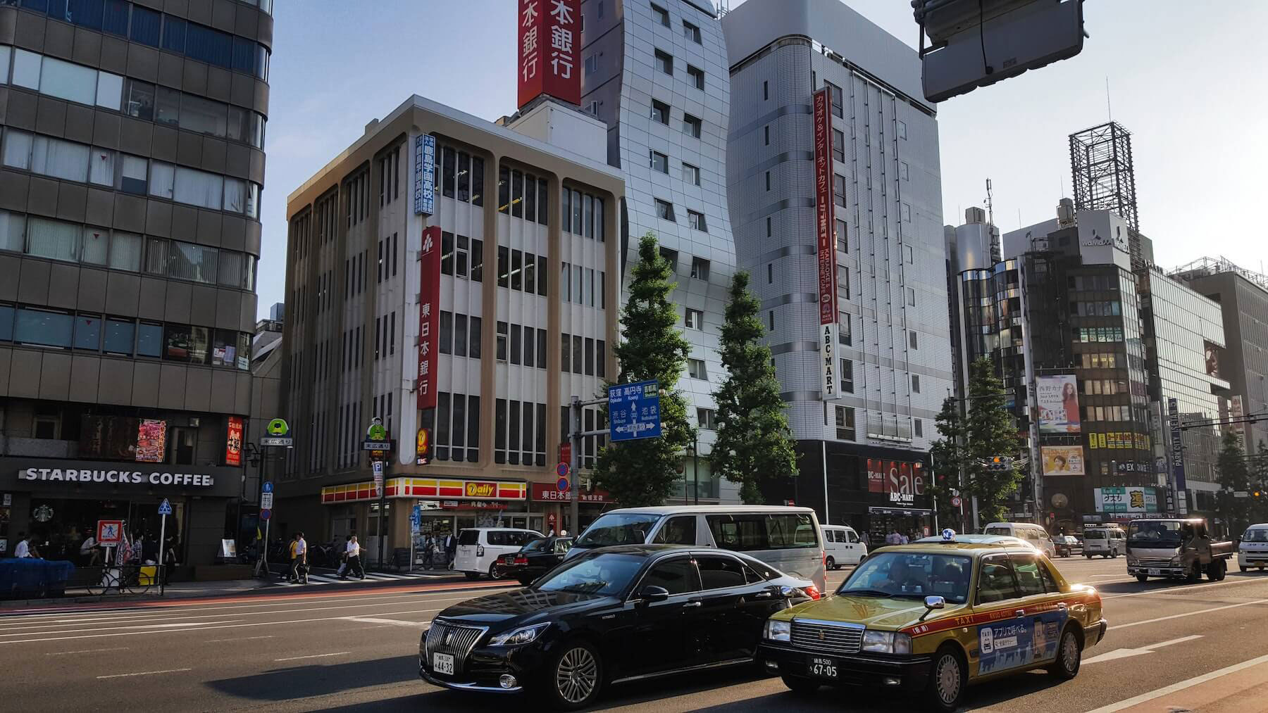The image shows a bustling city street with modern buildings, cars parked along the roadside, traffic lights, and pedestrians on the sidewalks.