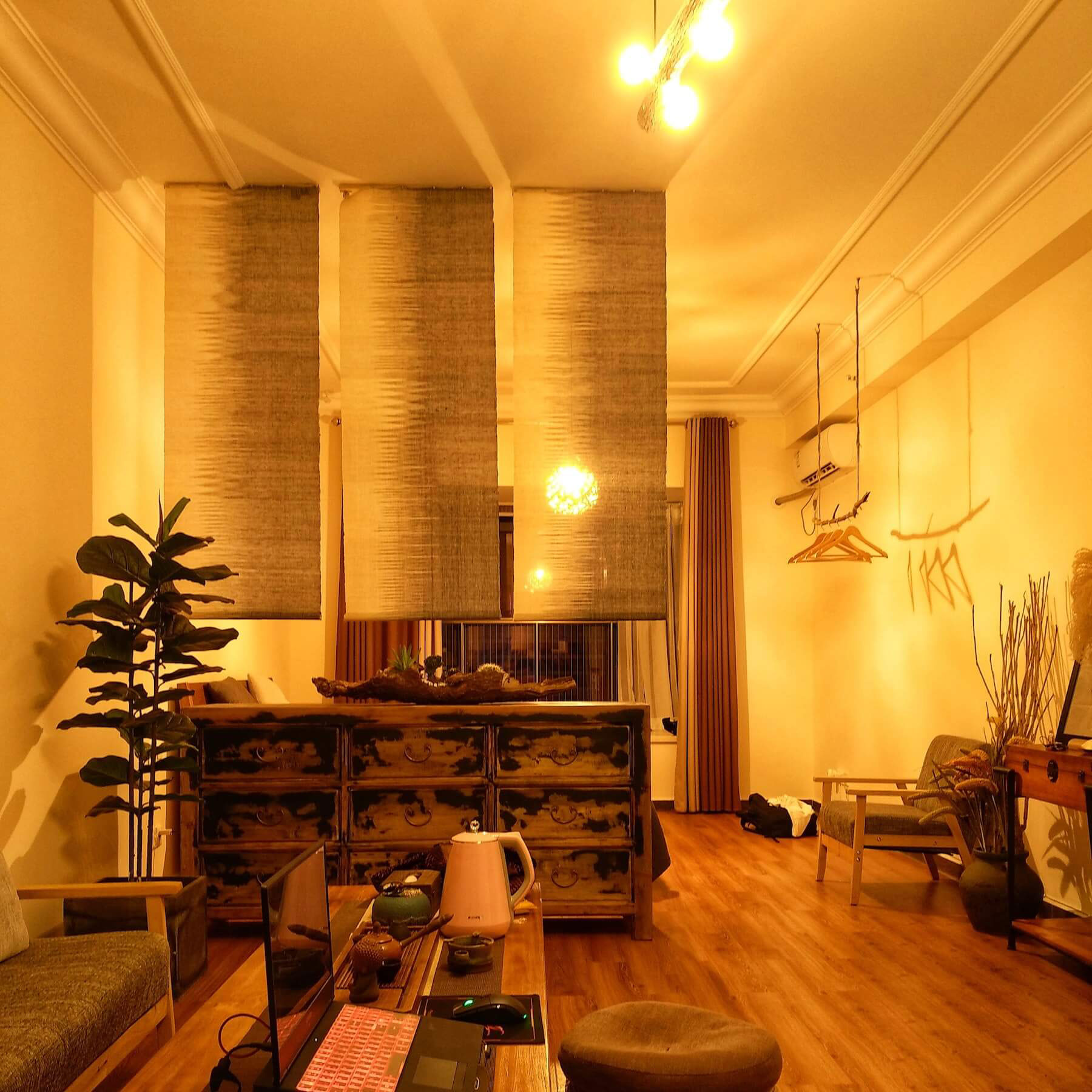 The image shows an interior view of a room with a high ceiling, exposed beams, hanging light fixtures, curtains, furniture, and decorative items, suggesting a contemporary living space.