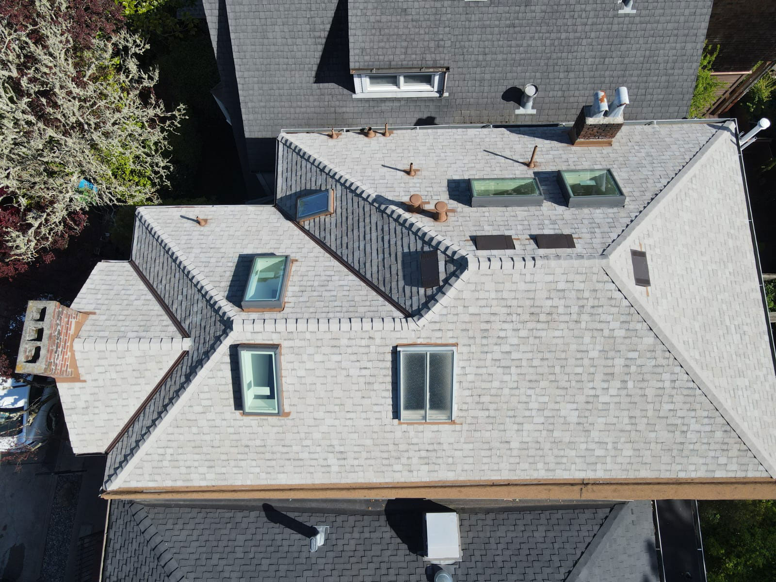 The image shows a modern residential building with a distinctive roof design featuring multiple sloped sections and flat areas, some of which have skylights and glass panels, set against a clear sky.