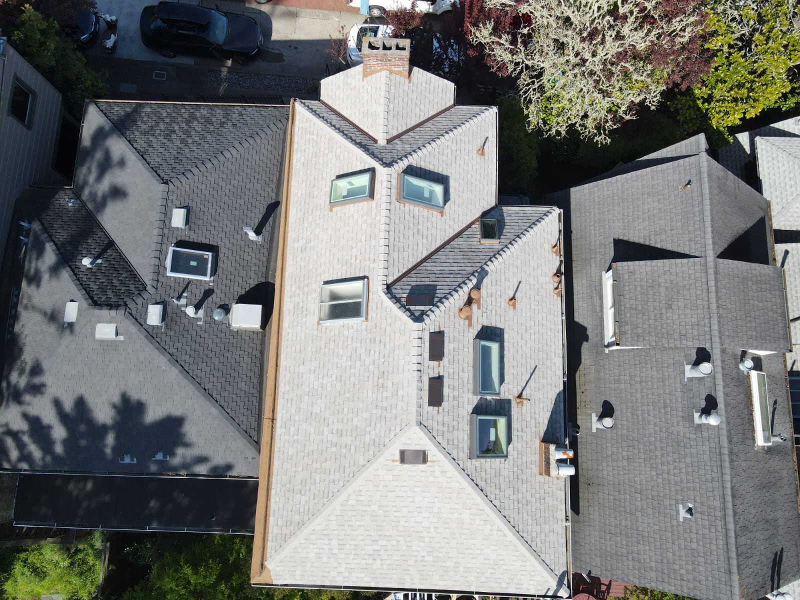 The image shows an elevated view of a residential house with a distinctive roof design featuring multiple sloped sections and a central skylight, set against a clear sky.