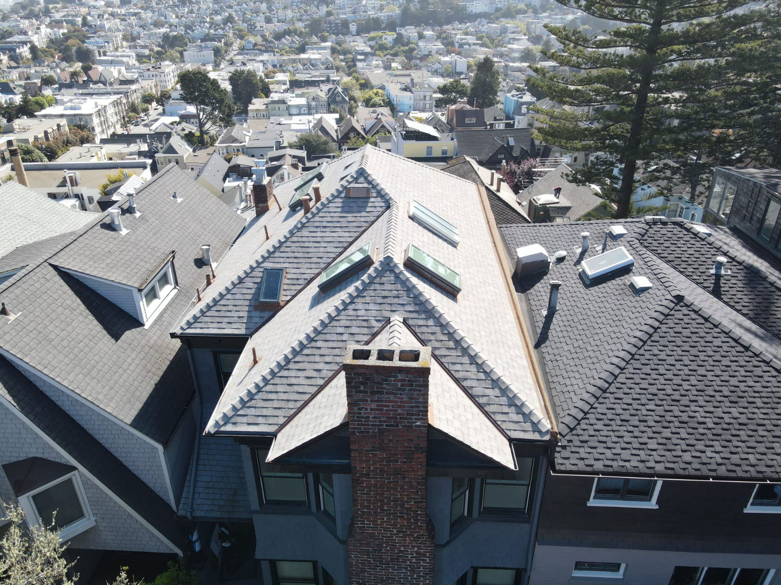 The image shows a multi-story residential building with a prominent roofline featuring multiple sloped sections and a central triangular feature, set against an urban background with other buildings and a clear sky.