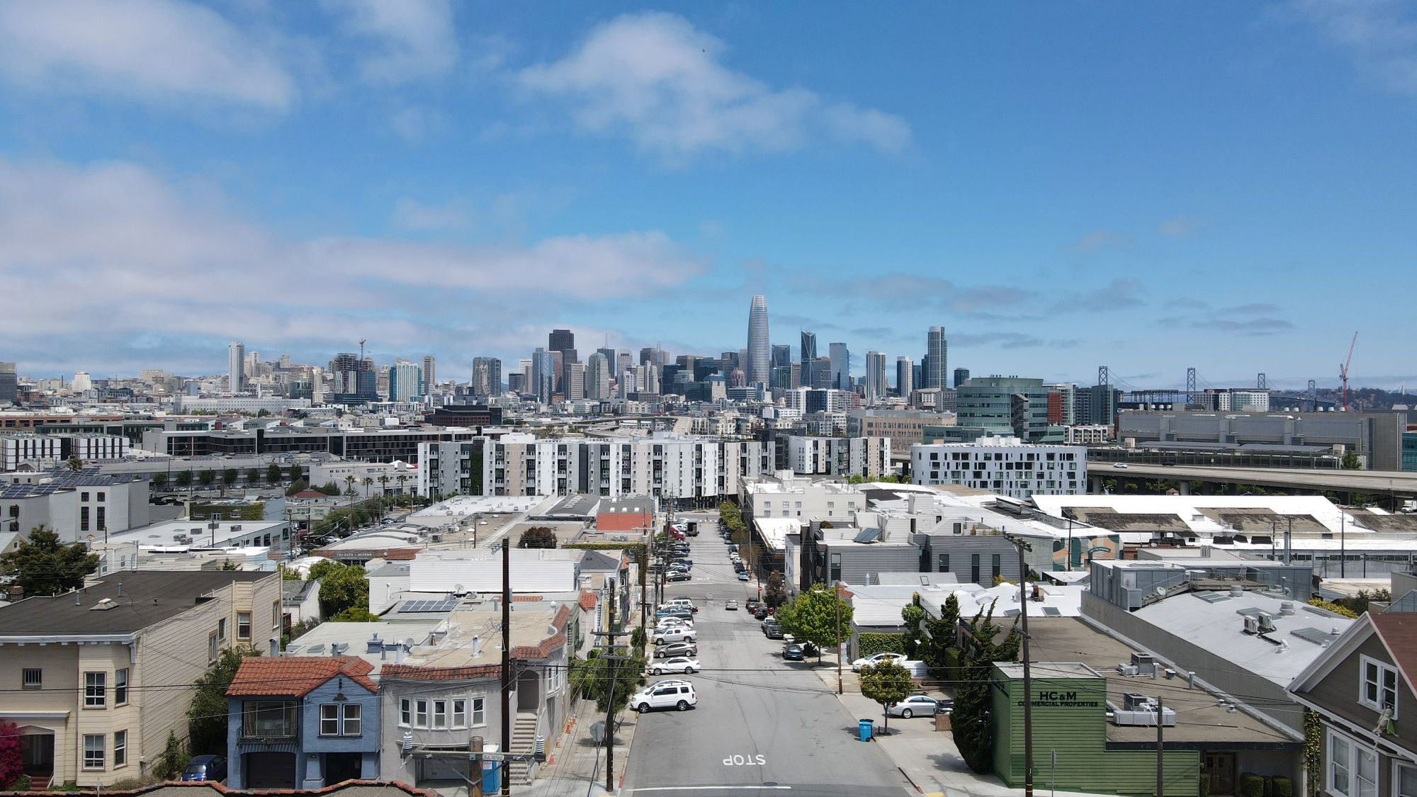 An urban scene with a city skyline under a clear blue sky, featuring a wide street lined with buildings and vehicles, set against a backdrop of a large body of water.