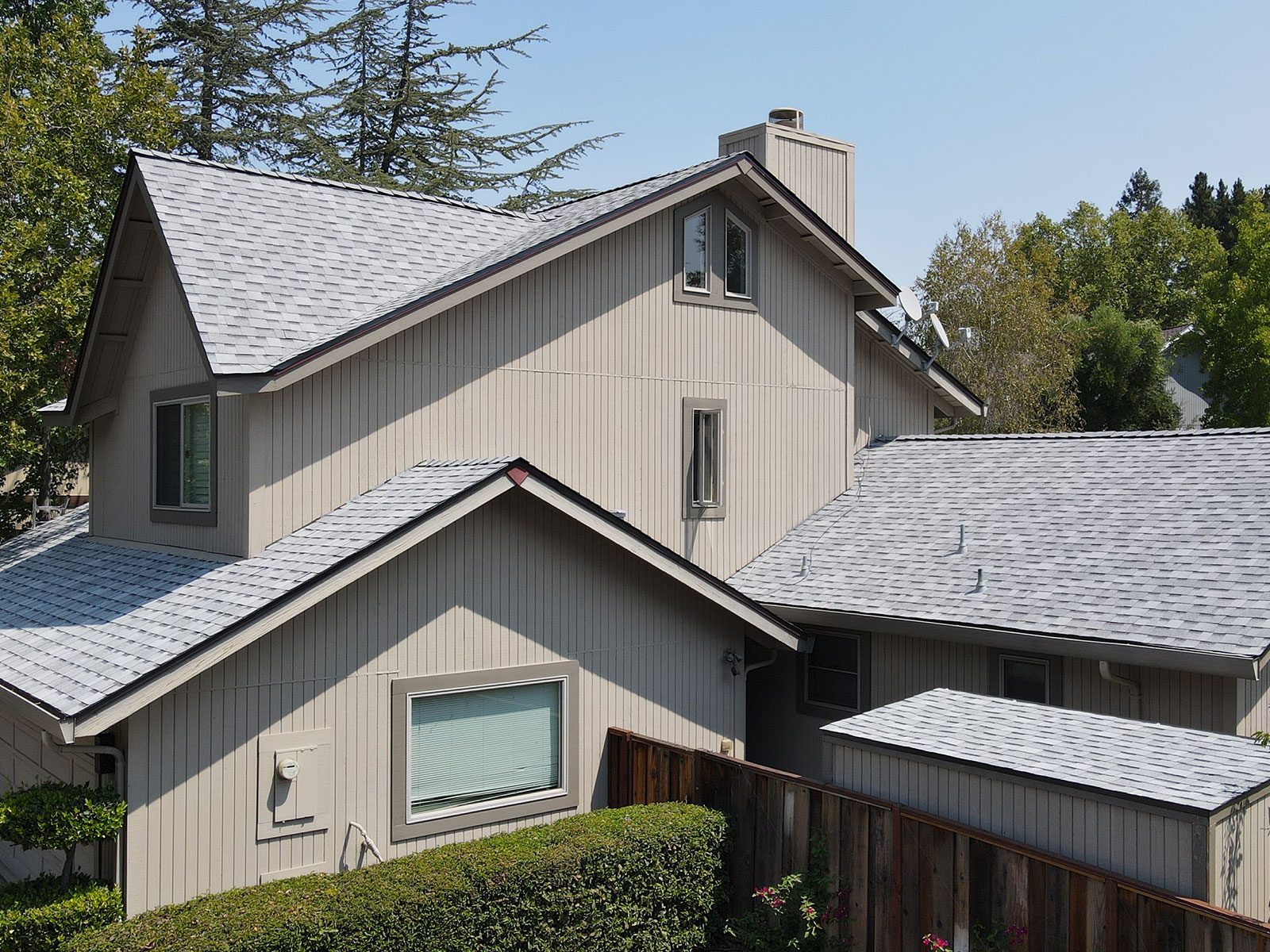 The image shows a two-story residential house with a gray shingle roof, featuring a combination of beige siding and darker trim around windows and doors, set against a clear sky on a sunny day.