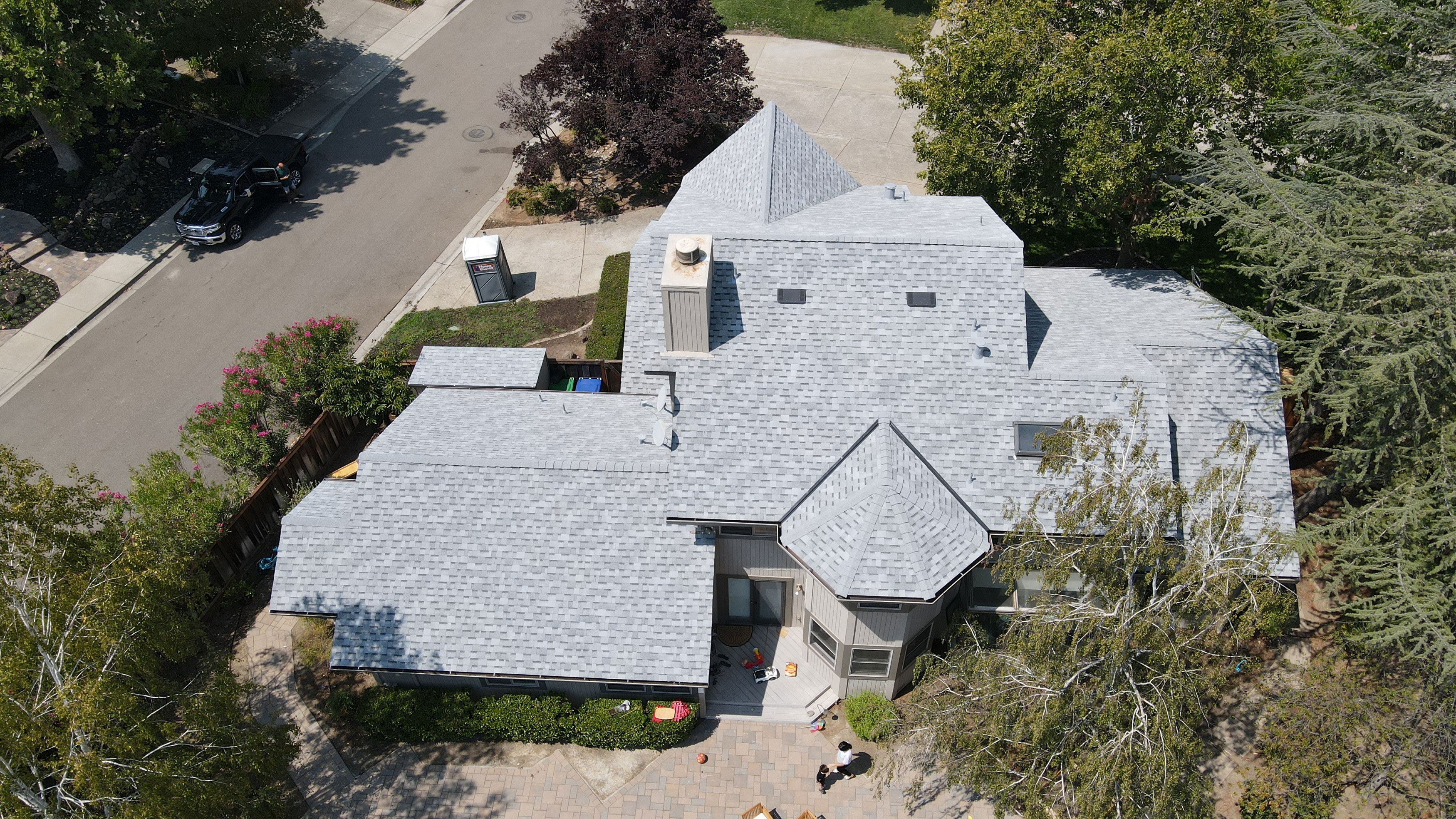 The image shows a two-story residential house with a gray roof and multiple windows and doors, set against a clear sky with trees visible in the background.