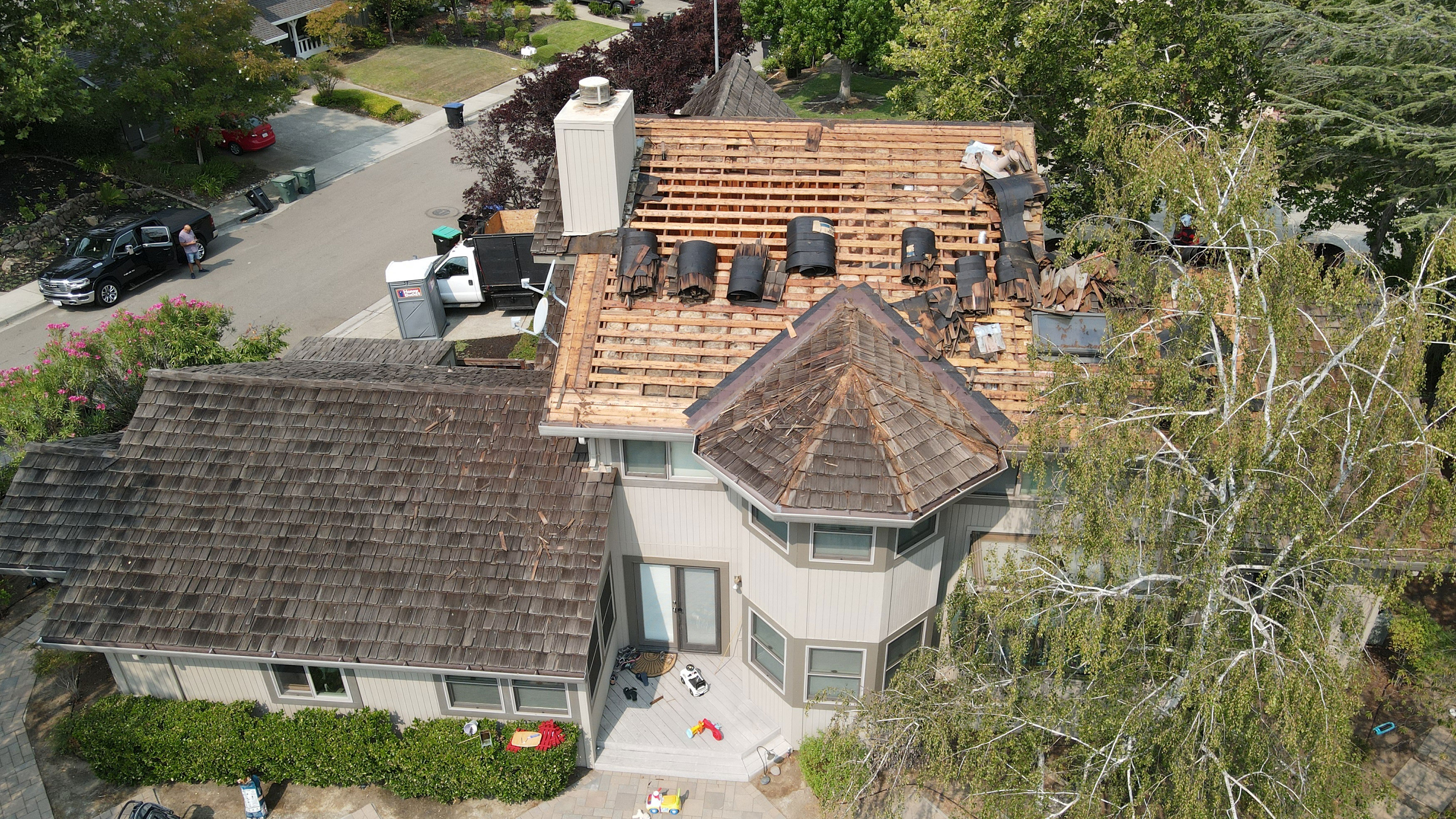 The image shows a two-story residential house with an unfinished roof and some shingles missing, undergoing construction or renovation.