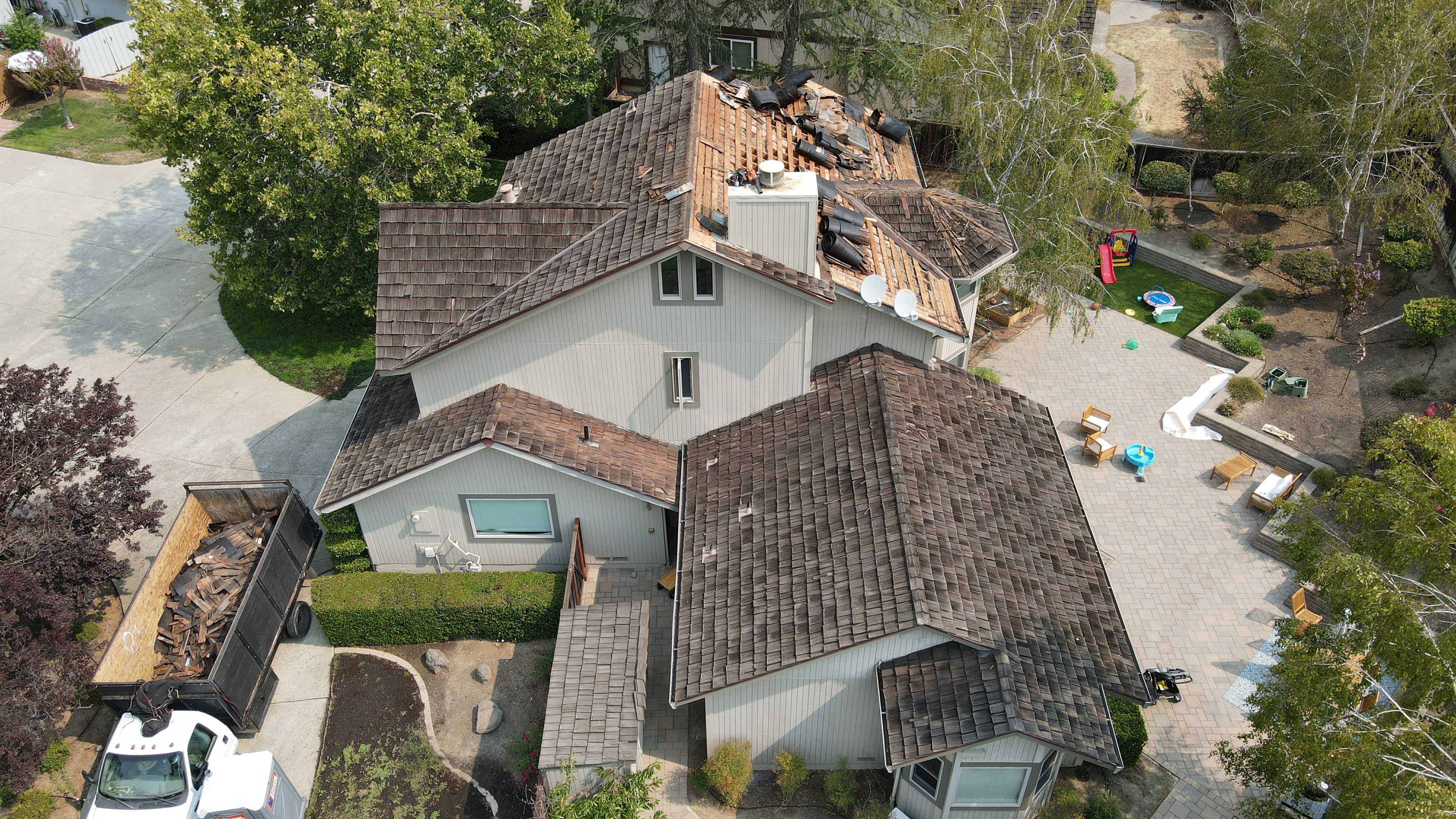 A two-story house with a pitched roof undergoing renovation, featuring exposed shingles and a partially covered front porch.