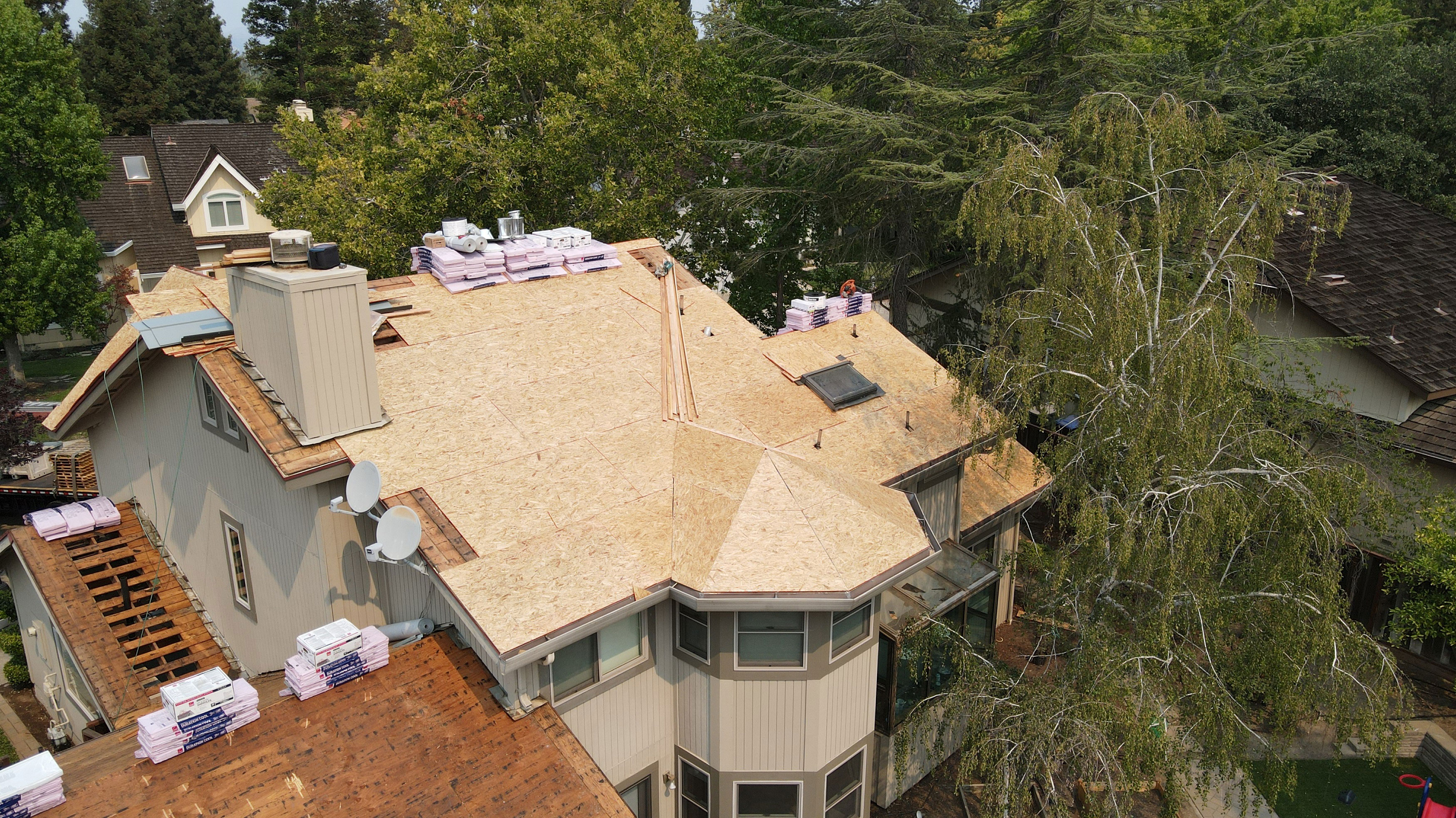 The image depicts a residential house undergoing roof renovation with visible construction materials such as plywood and insulation, indicating an ongoing remodeling project.
