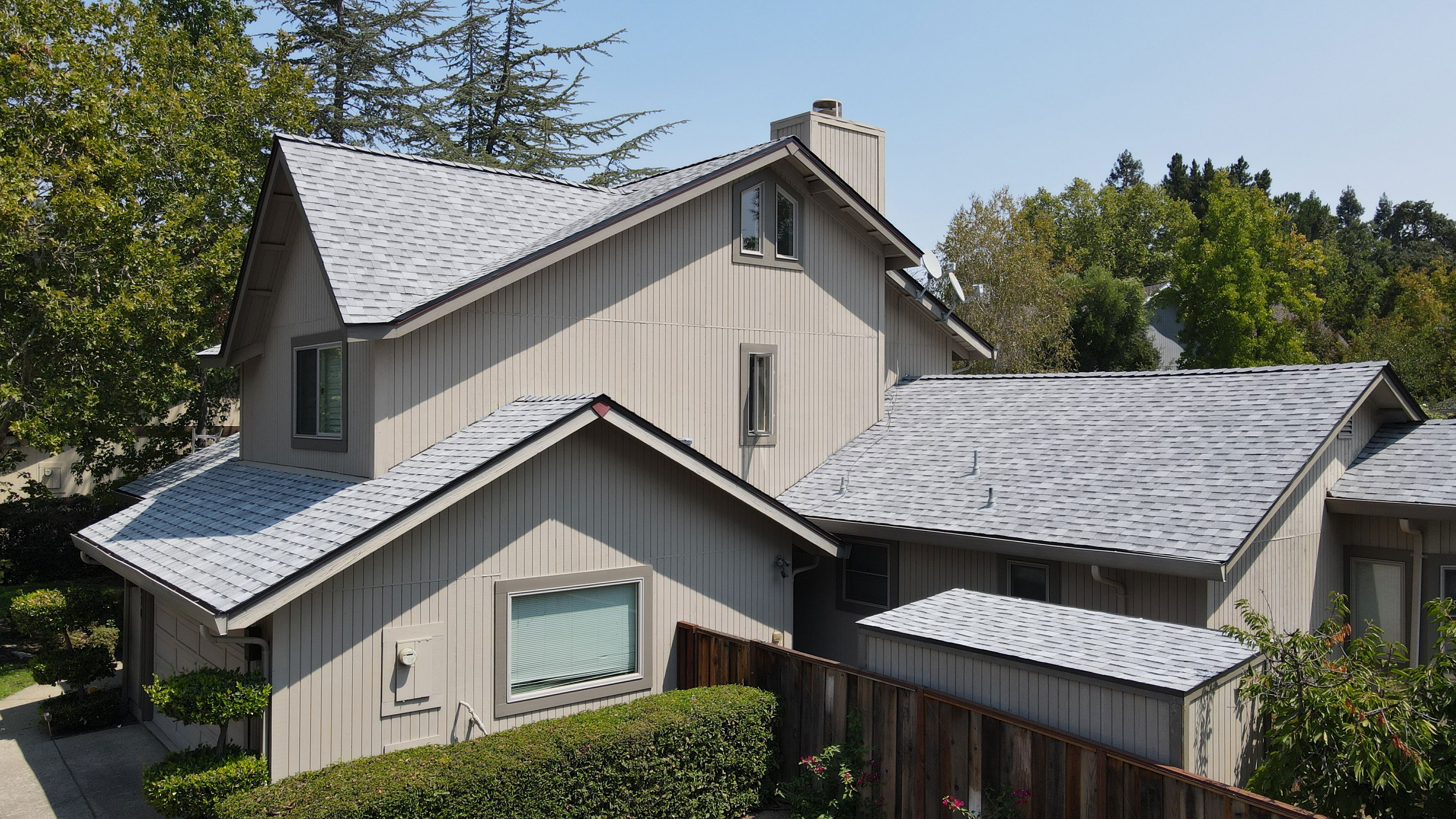 The image shows a two-story residential house with a pitched roof, grey siding, dark brown roof shingles, grey shutters, and a garage. There are multiple windows visible on both levels of the house, and the front yard features a well-maintained lawn and landscaping.
