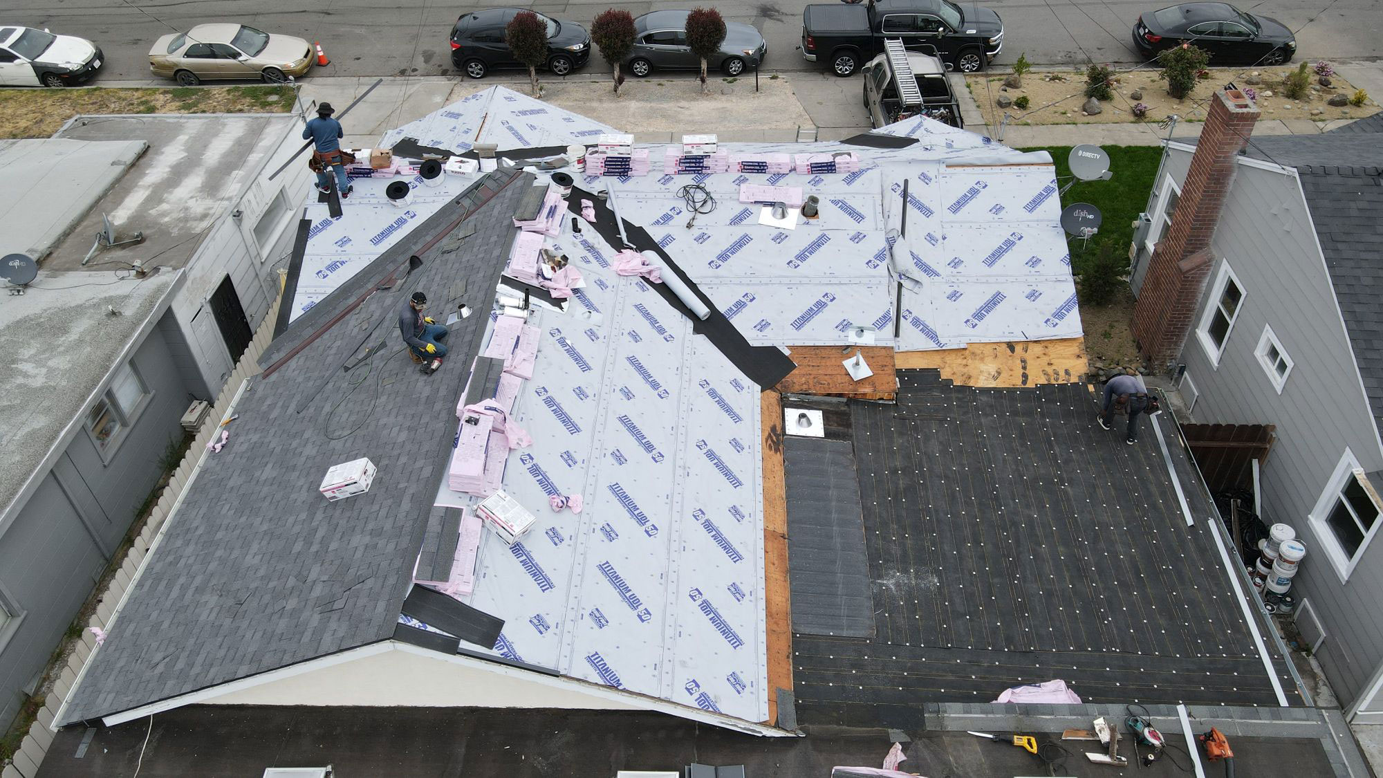 The image shows a residential building undergoing roofing construction, with workers visible on the scaffolding, and the surrounding area includes parked cars and a street view.