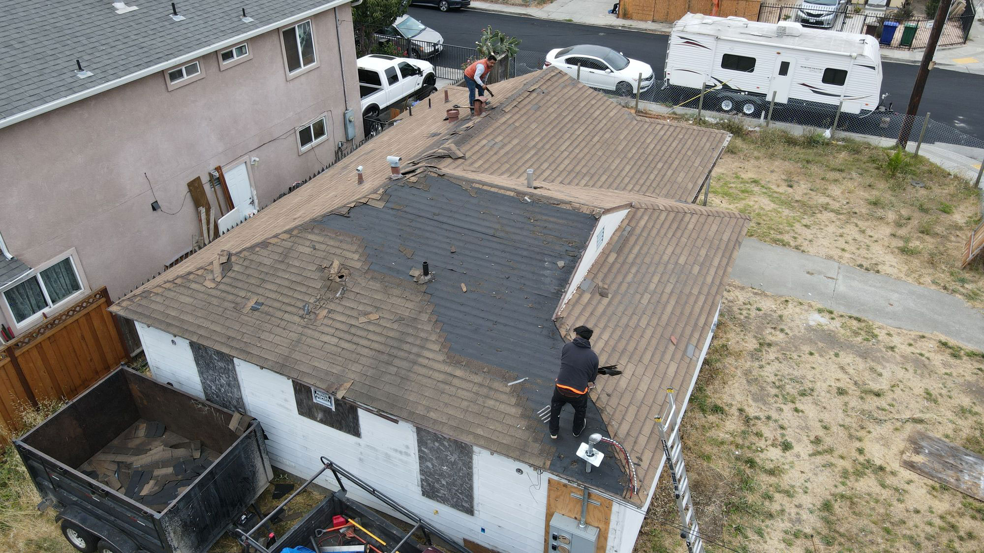 The image shows a residential house undergoing roof repair work with workers on the roof and ground, surrounded by building materials and tools.