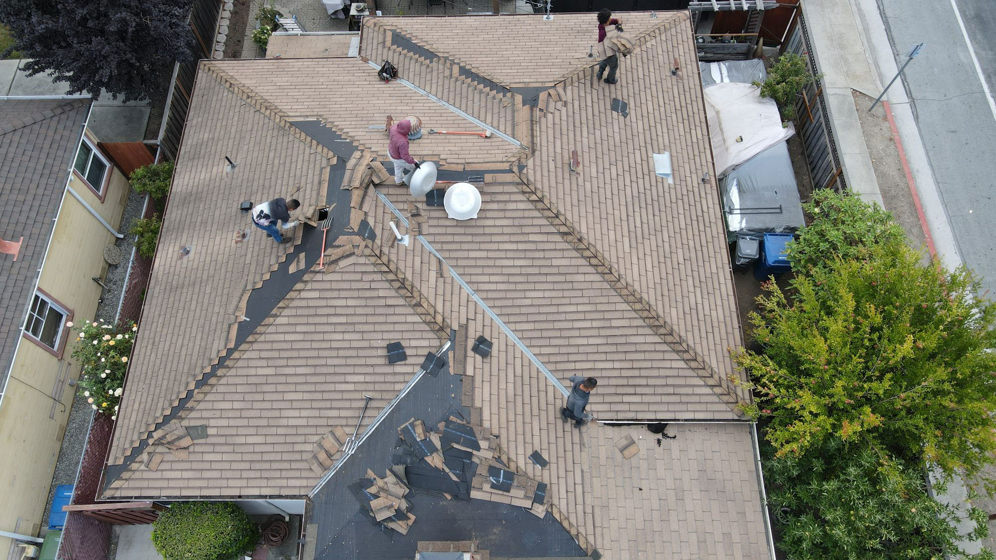 The image shows a house undergoing roof renovation with workers on the ground level and exposed rafters visible above them.