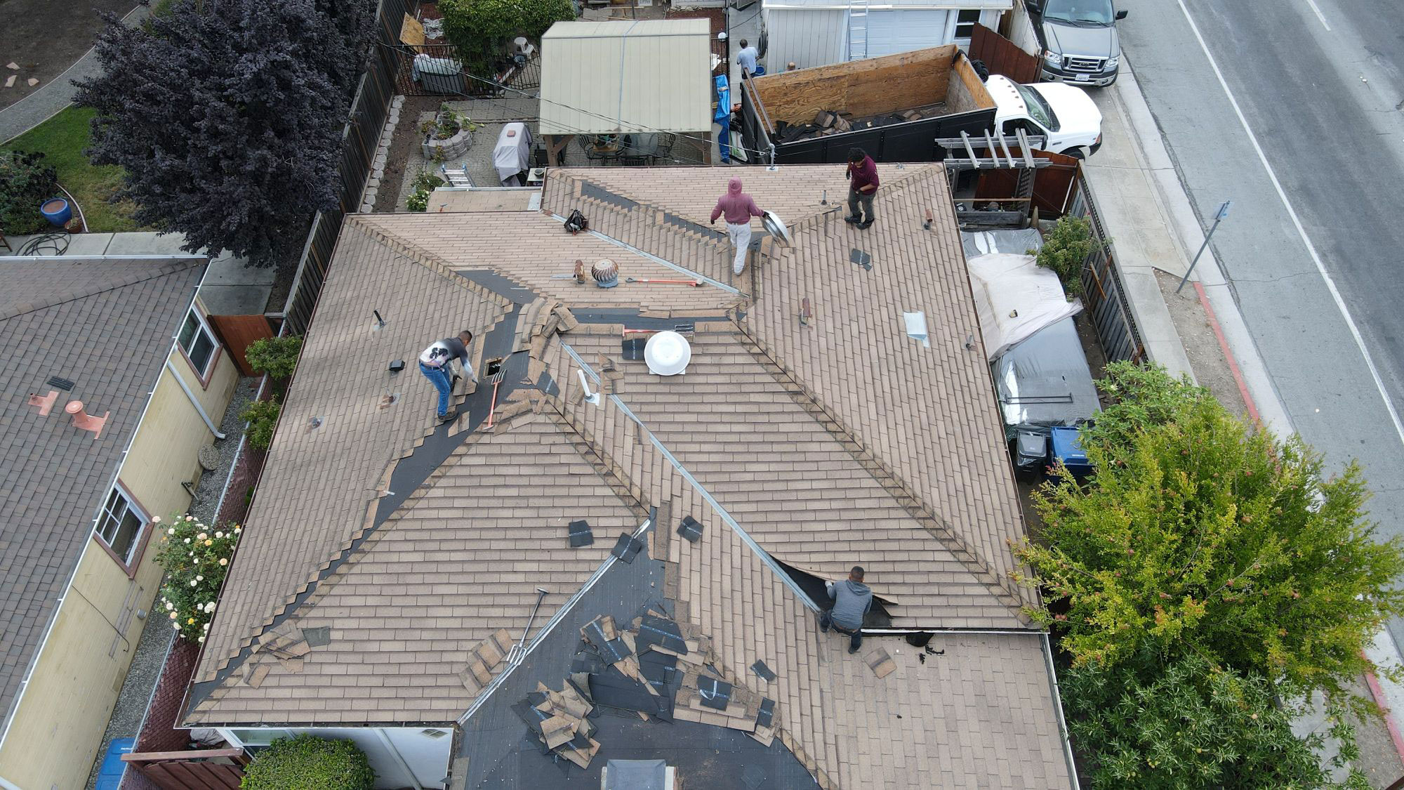 The image depicts a residential property with a damaged roof under repair, featuring workers on the ground and exposed wooden decking above.