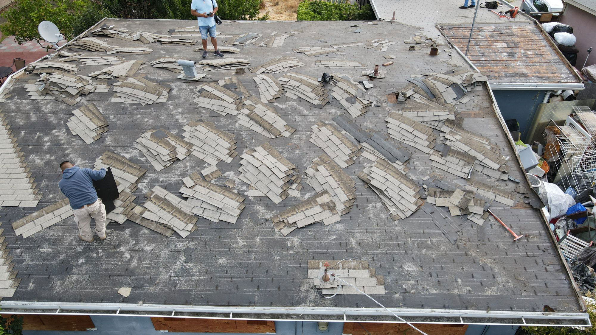 The image shows a roof with damaged shingles under construction, with workers on the ground and debris scattered around.