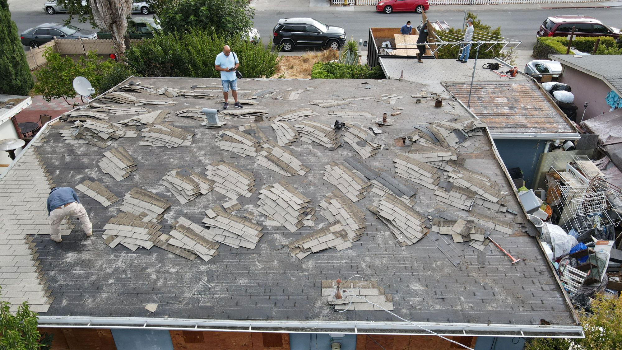 The image shows a person standing on a rooftop with damaged shingles, surrounded by debris, indicating ongoing roof repair work.