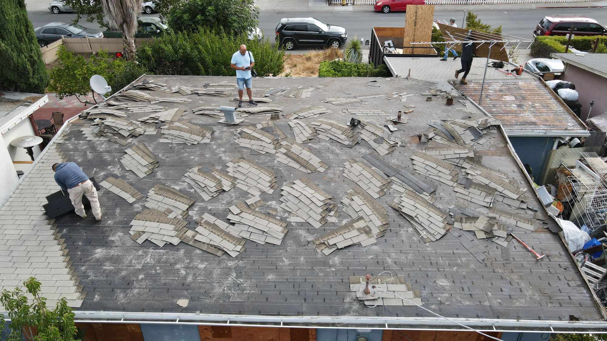 The image shows a rooftop undergoing repair work with visible shingles removed and being replaced, workers on the roof, and the surrounding area appears to be an urban environment with houses and cars.
