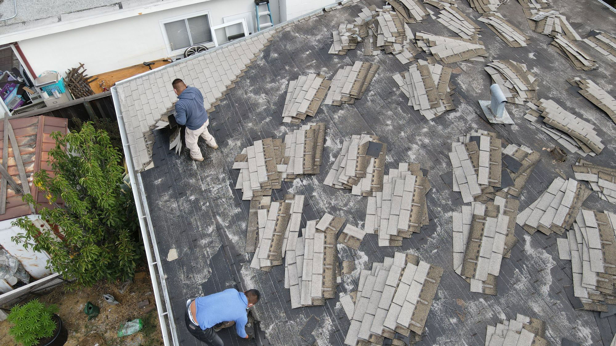 The image shows a person working on a roof with shingles being replaced or removed, indicating maintenance or renovation activity.