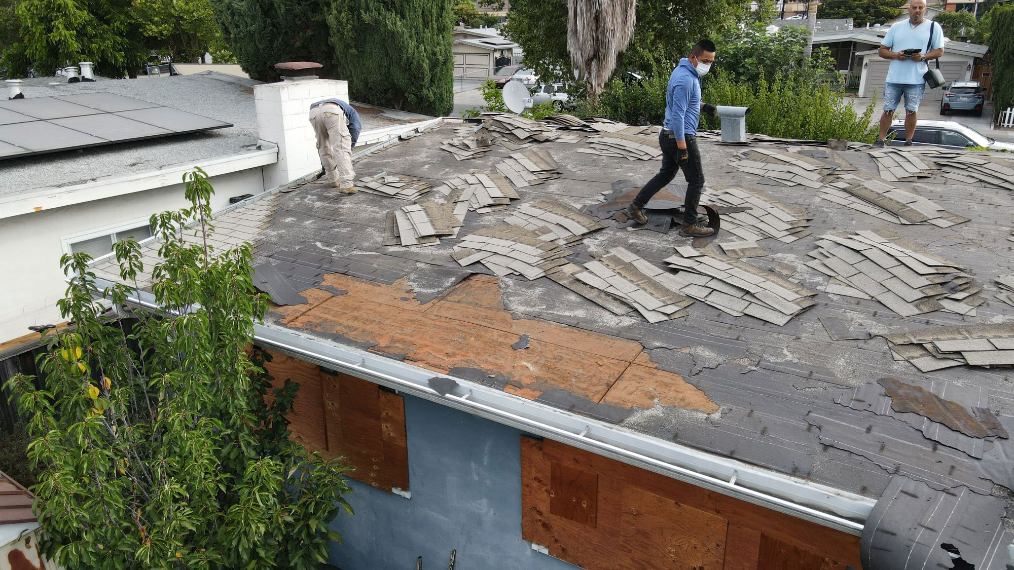 This is a photograph showing a person working on a roof with damaged shingles, surrounded by a residential area with other houses visible in the background.
