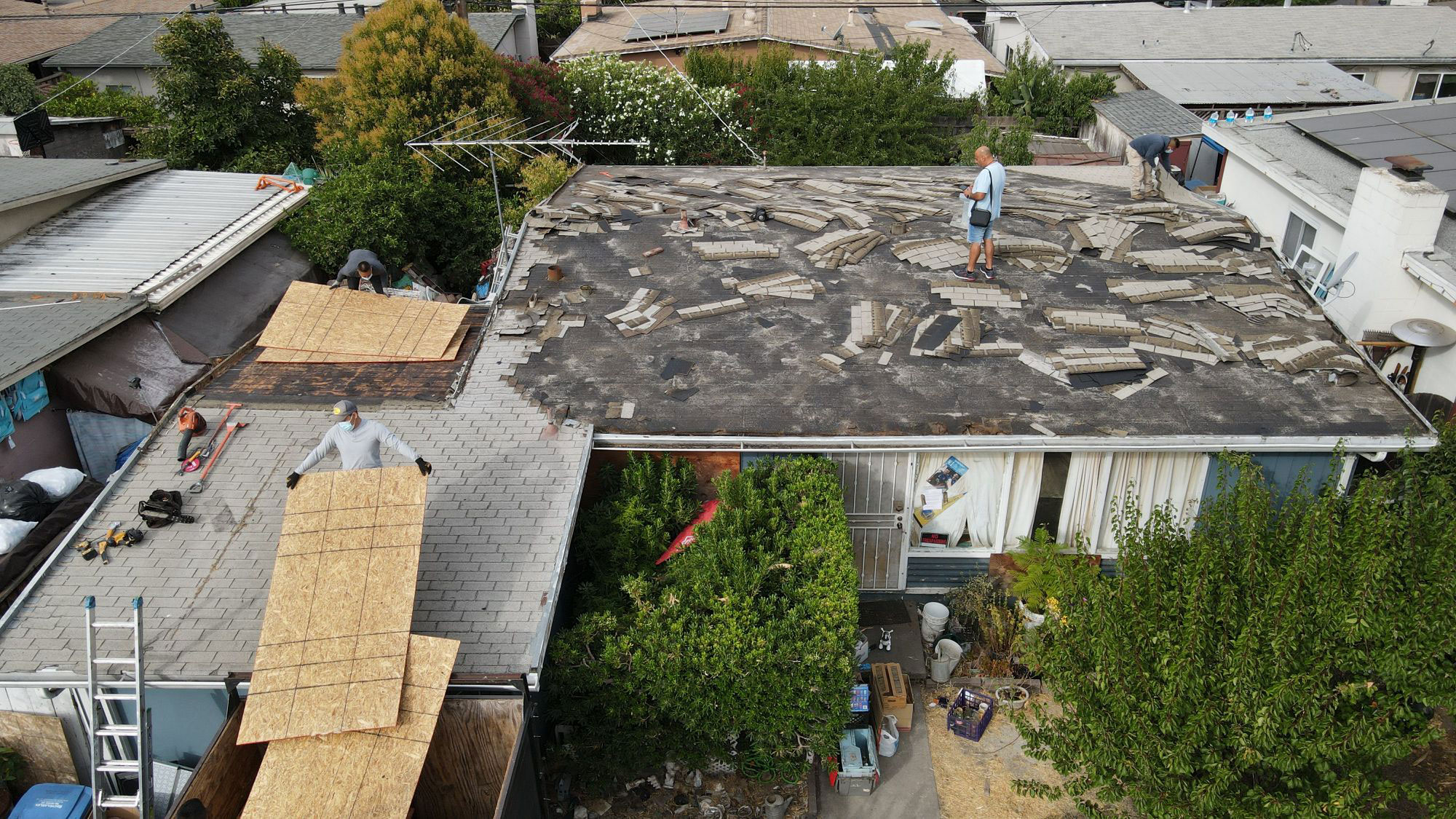 The image shows a residential area with a house undergoing roof repairs. A person is standing on the roof amidst construction materials, indicating ongoing work.