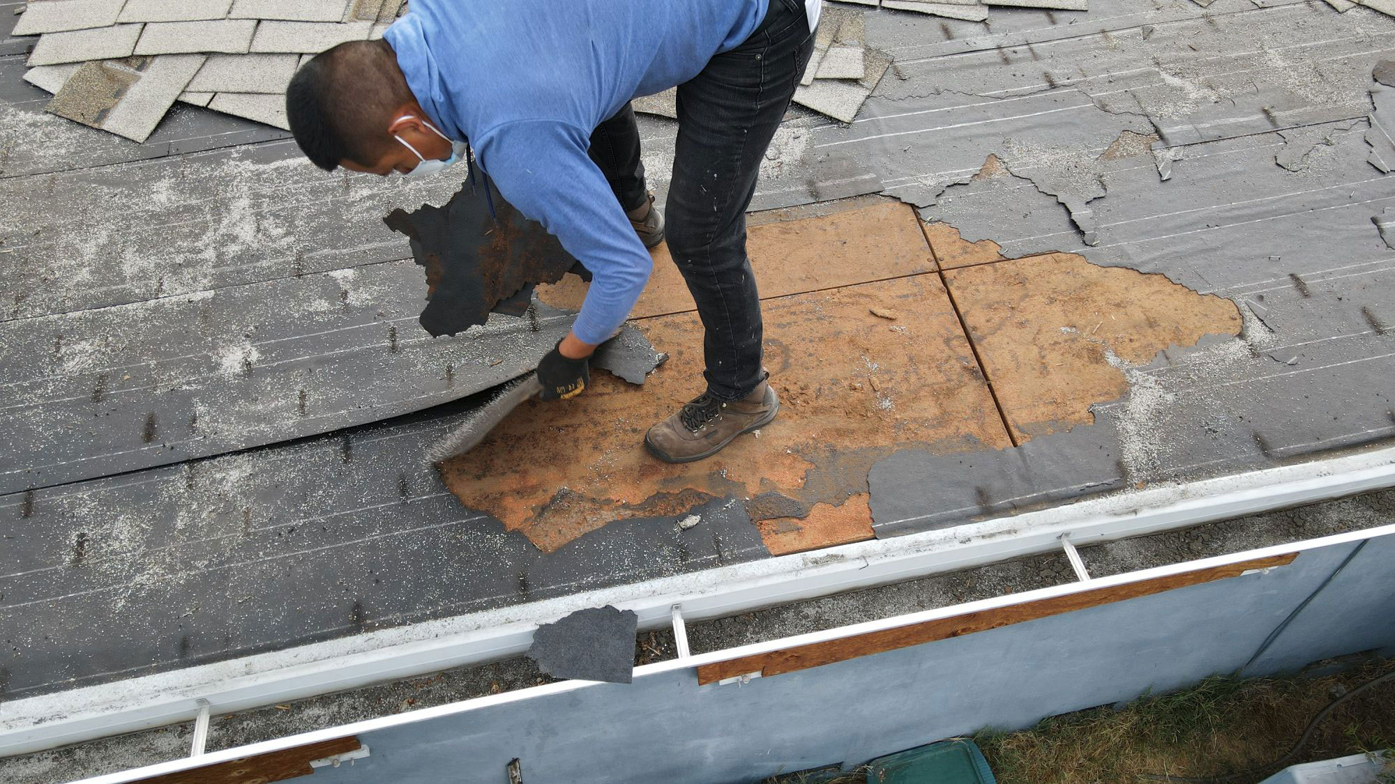 The image depicts a person working on a damaged roof, with shingles removed and exposed plywood beneath.