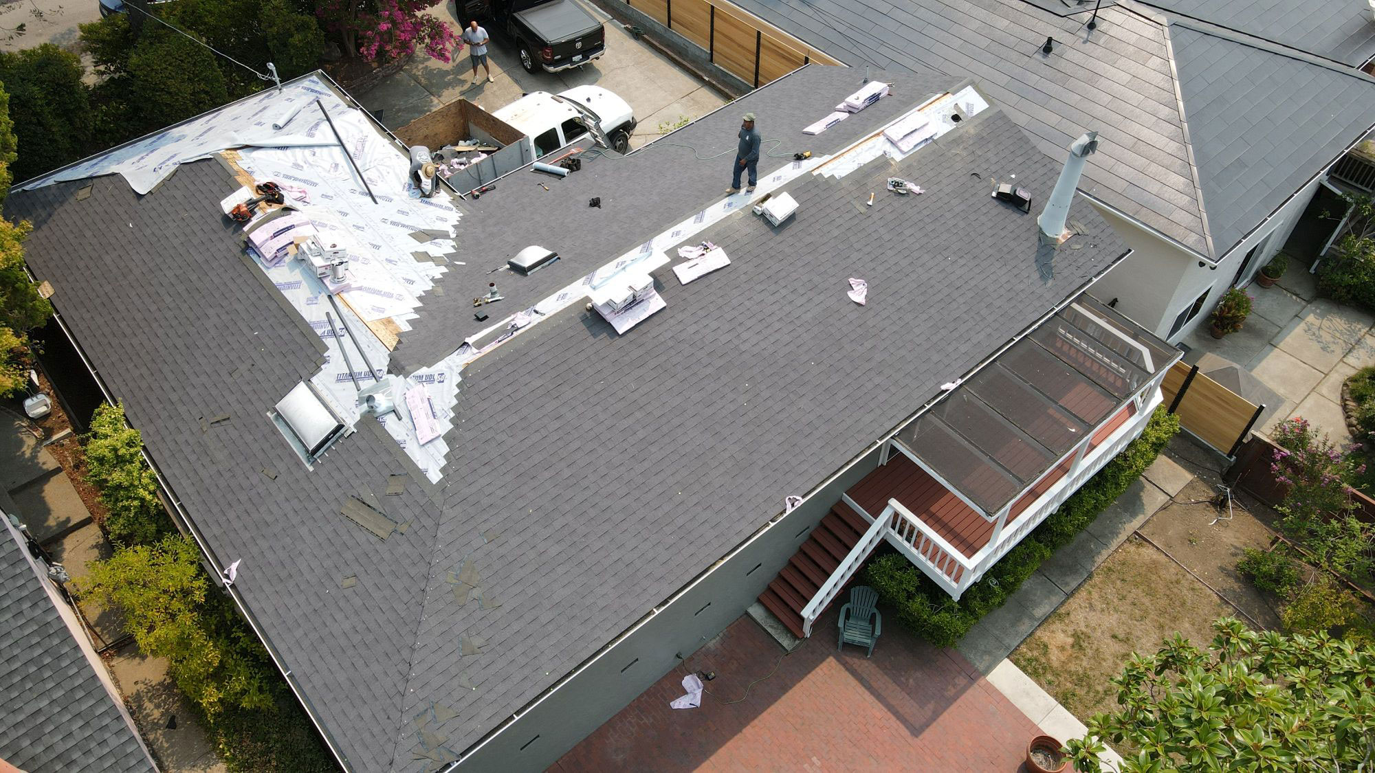 The image shows a house with a roof under repair, featuring exposed plywood and shingles being replaced.