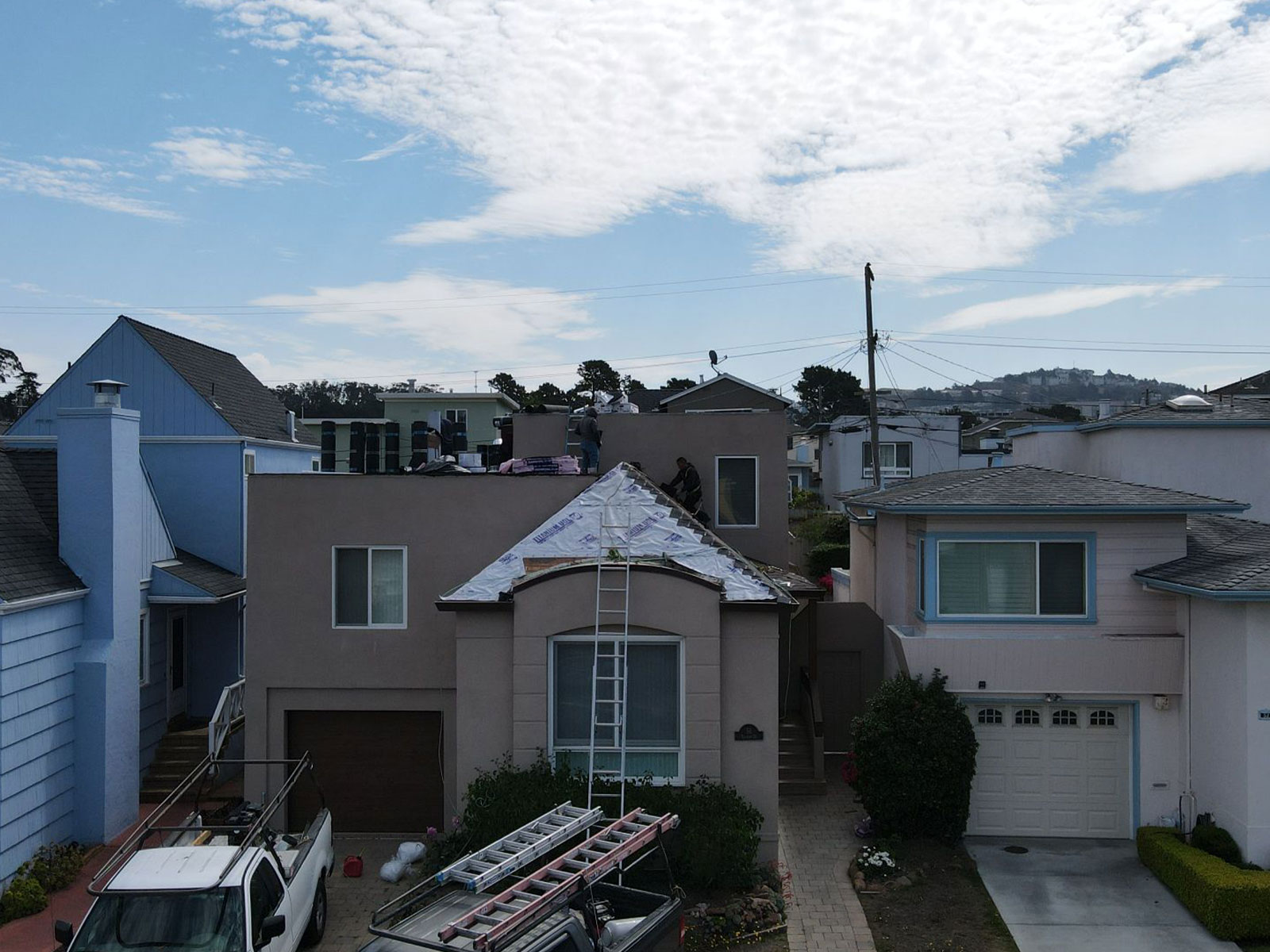 The image shows a residential street with two-story houses on either side, one of which has a visible roof repair underway.