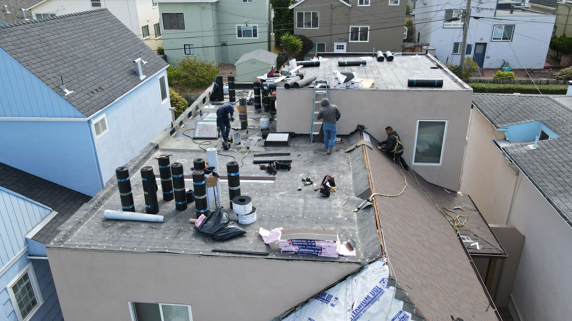The image shows a construction site with workers at the roof level of a building under renovation, featuring exposed insulation, plywood, and various materials scattered around.