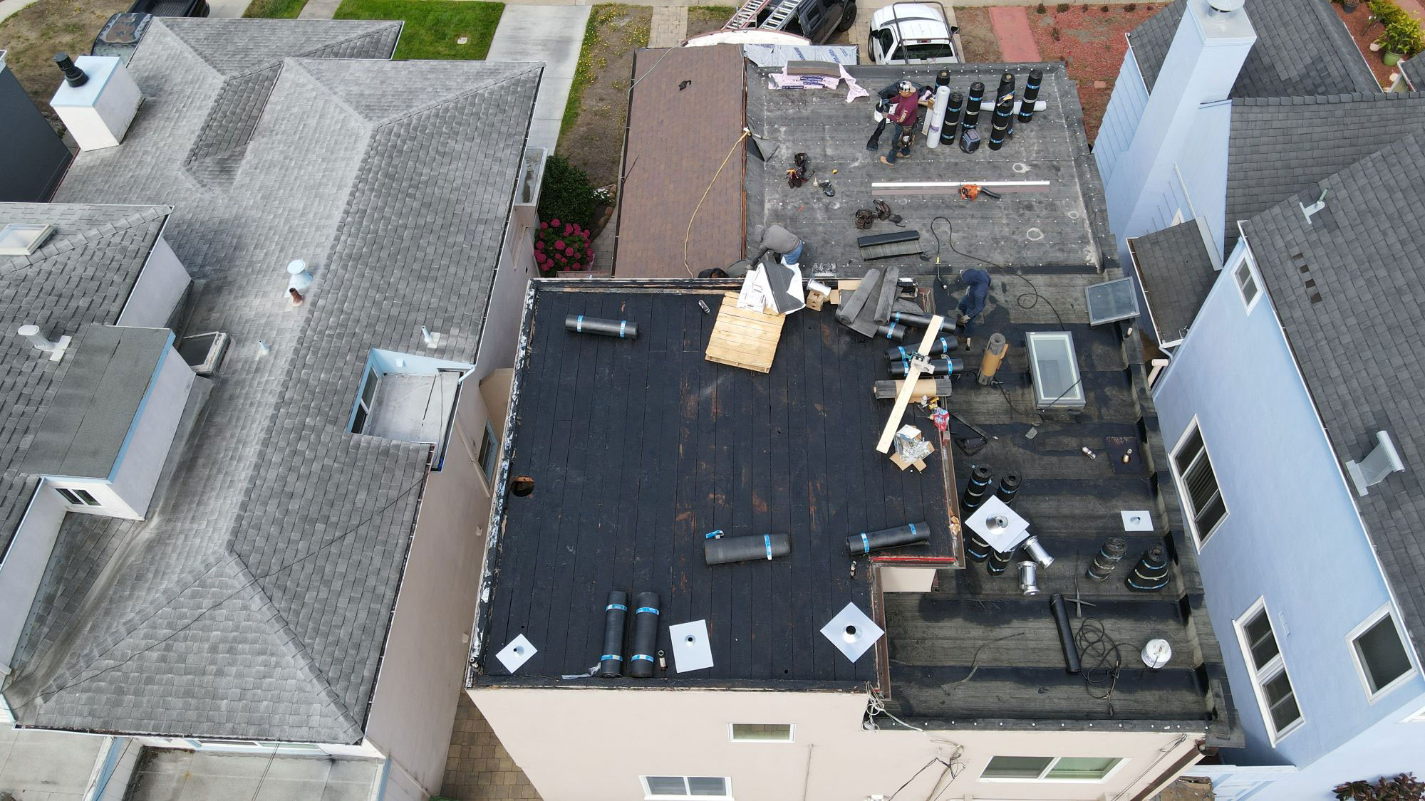 The image shows a residential building with a flat roof under construction, featuring exposed rafters and materials, alongside an adjacent house with a completed roof.