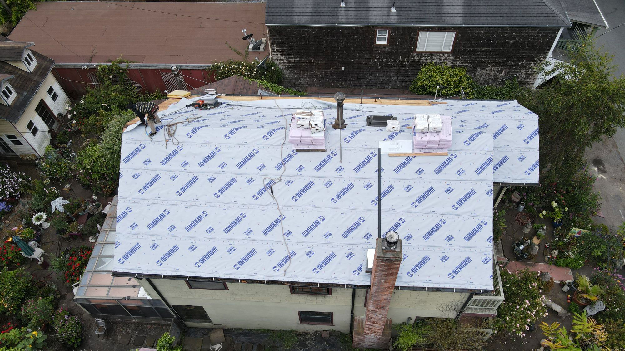 The image shows a house under construction with a visible roofing project in progress, featuring tarps and protective coverings.