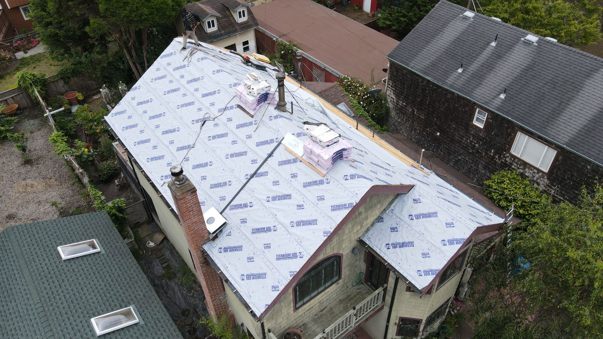 The image shows a residential house with a roof under construction, featuring new shingles, visible insulation, and a plastic tarp covering part of the structure.