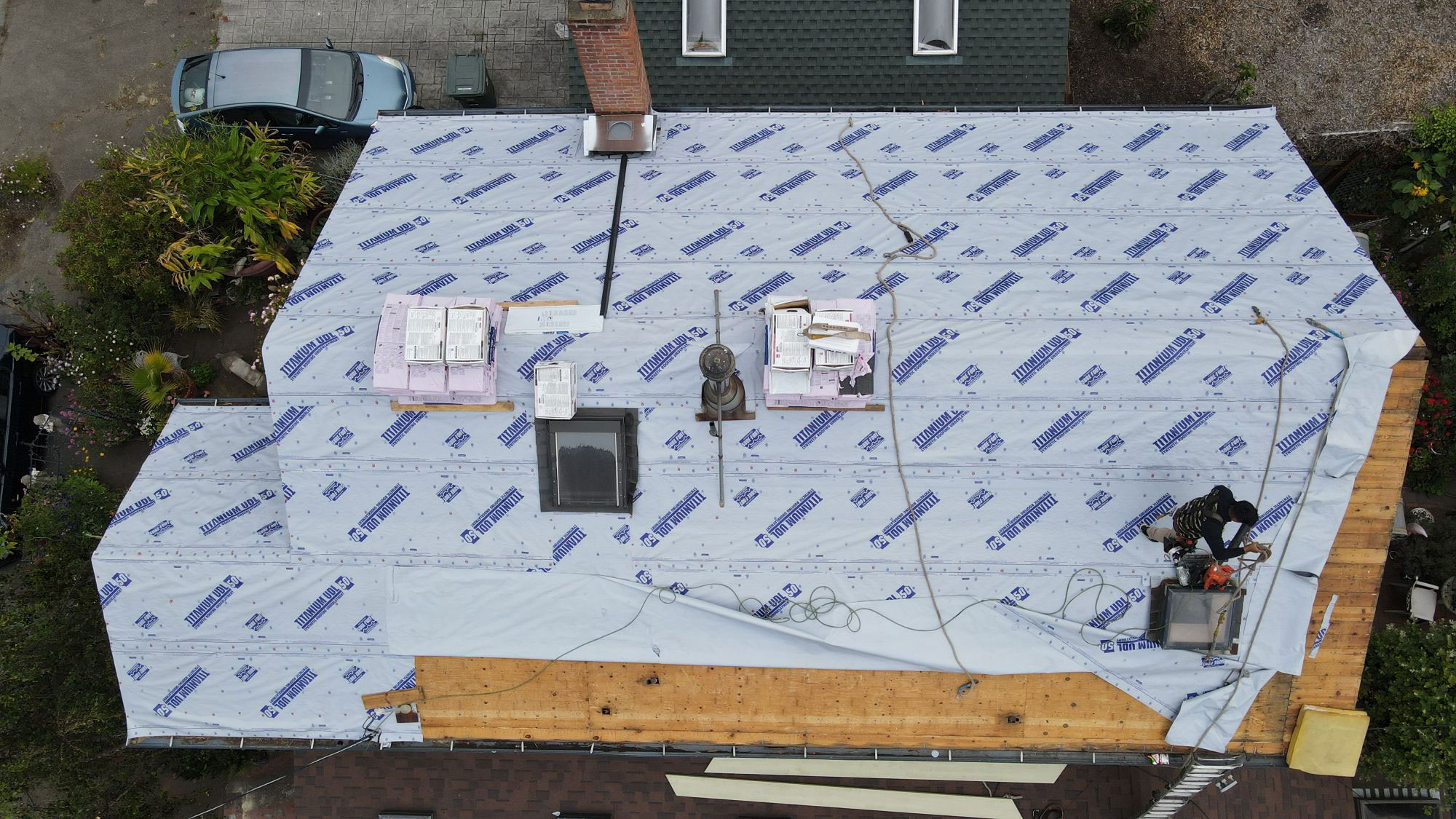 The image shows a house undergoing construction with a tarp covering the roof and some building materials visible on the ground.