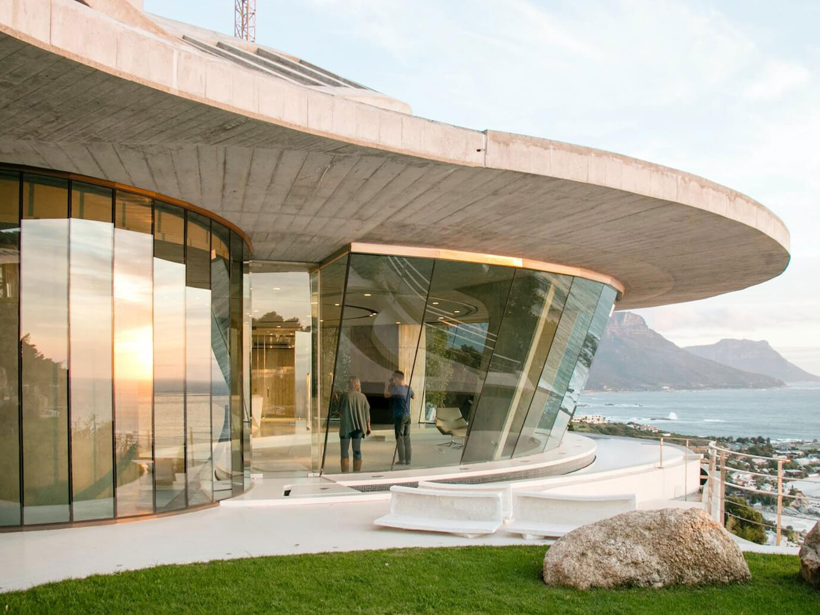 The image shows a modern architectural structure with curved glass walls, possibly a building entrance or lobby, situated near a body of water with a mountainous backdrop, under a clear sky during what appears to be either sunrise or sunset.