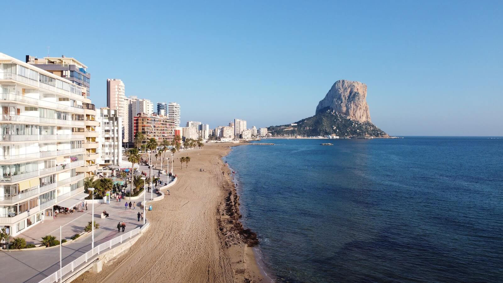 The image shows a scenic coastal view with a clear blue sky, a large rock formation resembling a castle, known as the Rock of Gibraltar, in the background, and a sandy beach with buildings and people in the foreground.