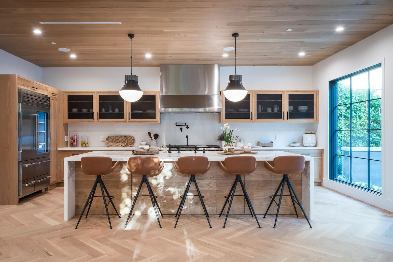 A modern kitchen with light wood cabinets, white countertops, black stools, metal appliances, and a large island in the center.