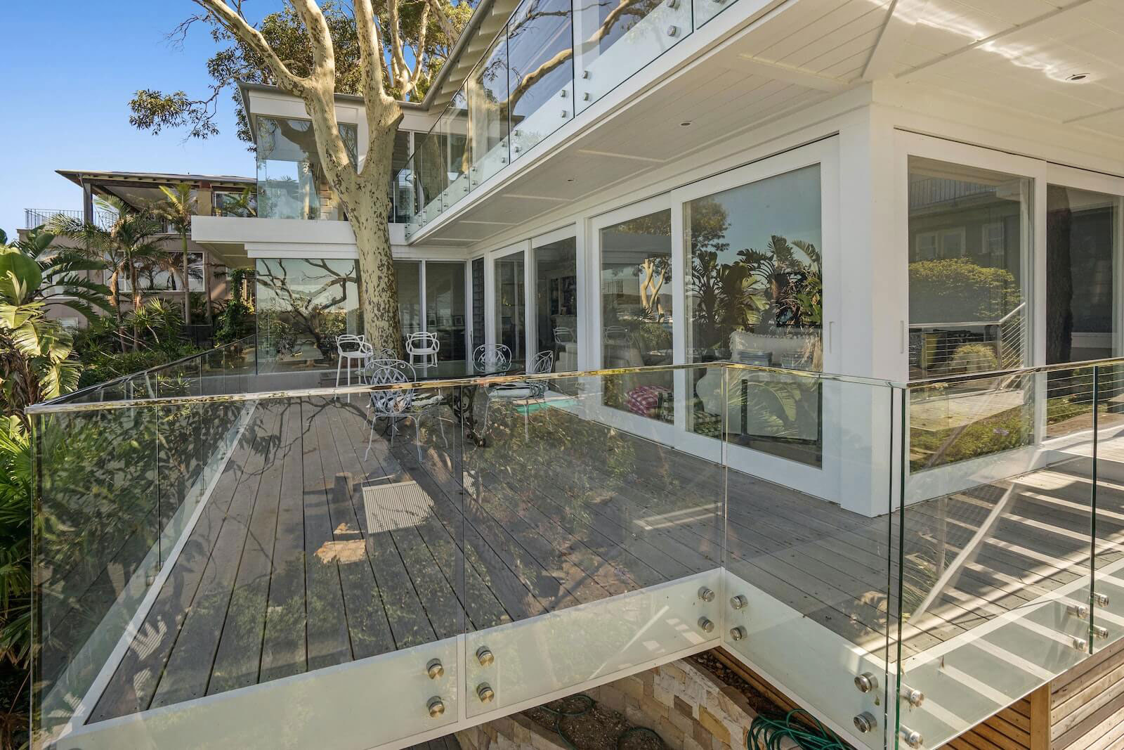 The image shows a modern two-story house with a glass railing on an upper deck overlooking a pool area, featuring a clear blue sky above.