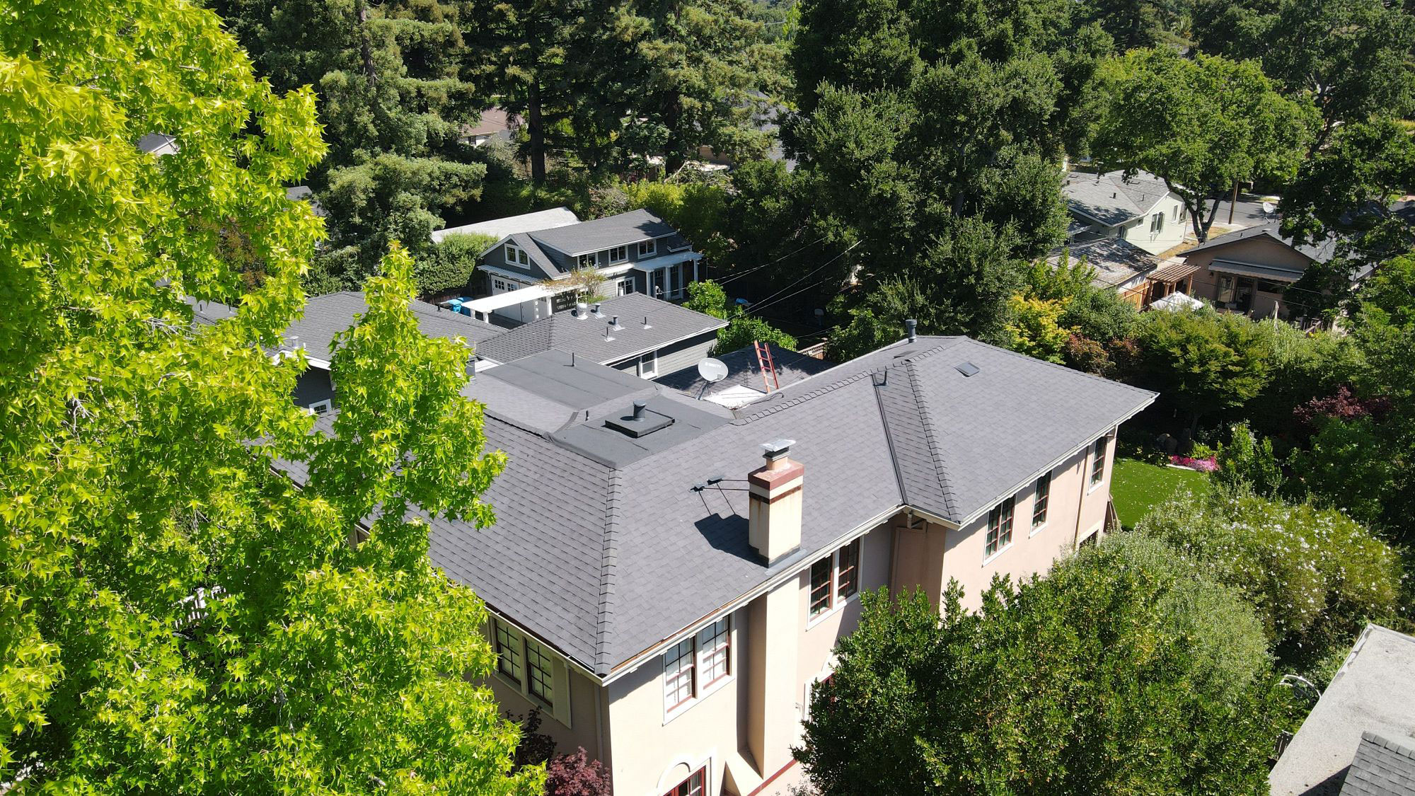 The image shows a two-story residential house with a gray roof, situated on a tree-lined street during the daytime under clear skies.