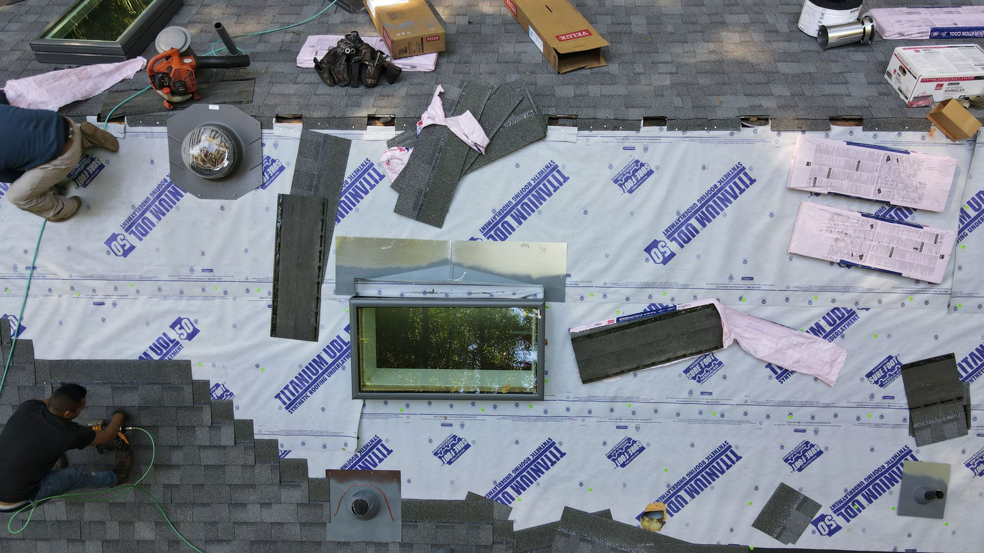 The image shows a person working on a roof under construction, with materials such as tarps and insulation scattered around, indicating an ongoing home renovation project.