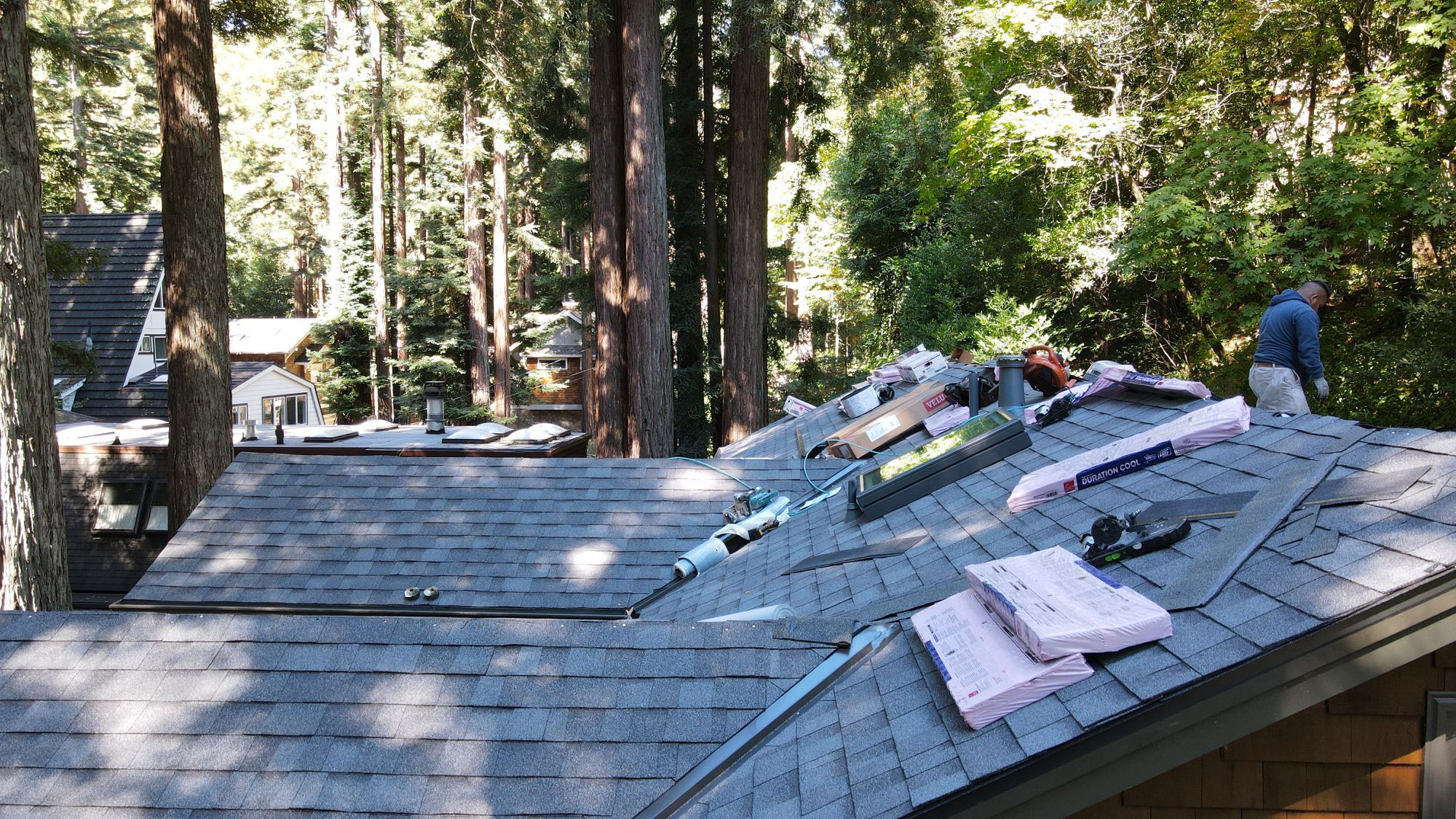 The image displays a house with an unfinished roof under construction, featuring exposed rafters and shingles, taken during daylight with natural surroundings.