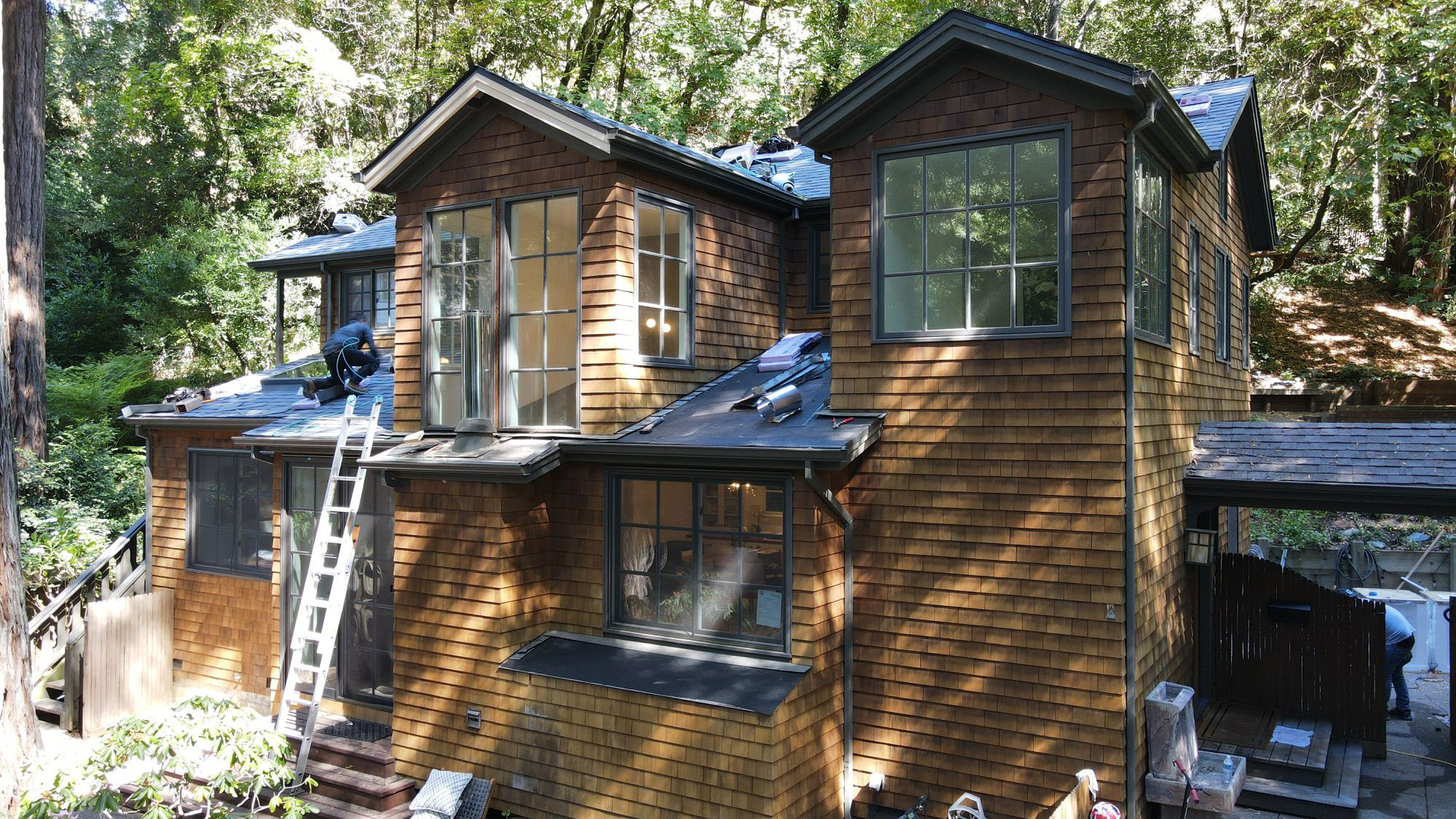 The image shows a two-story residential house with a dark roof, visible solar panels, and a wooden deck. It appears to be under construction or renovation, with some areas covered by scaffolding or protective coverings.