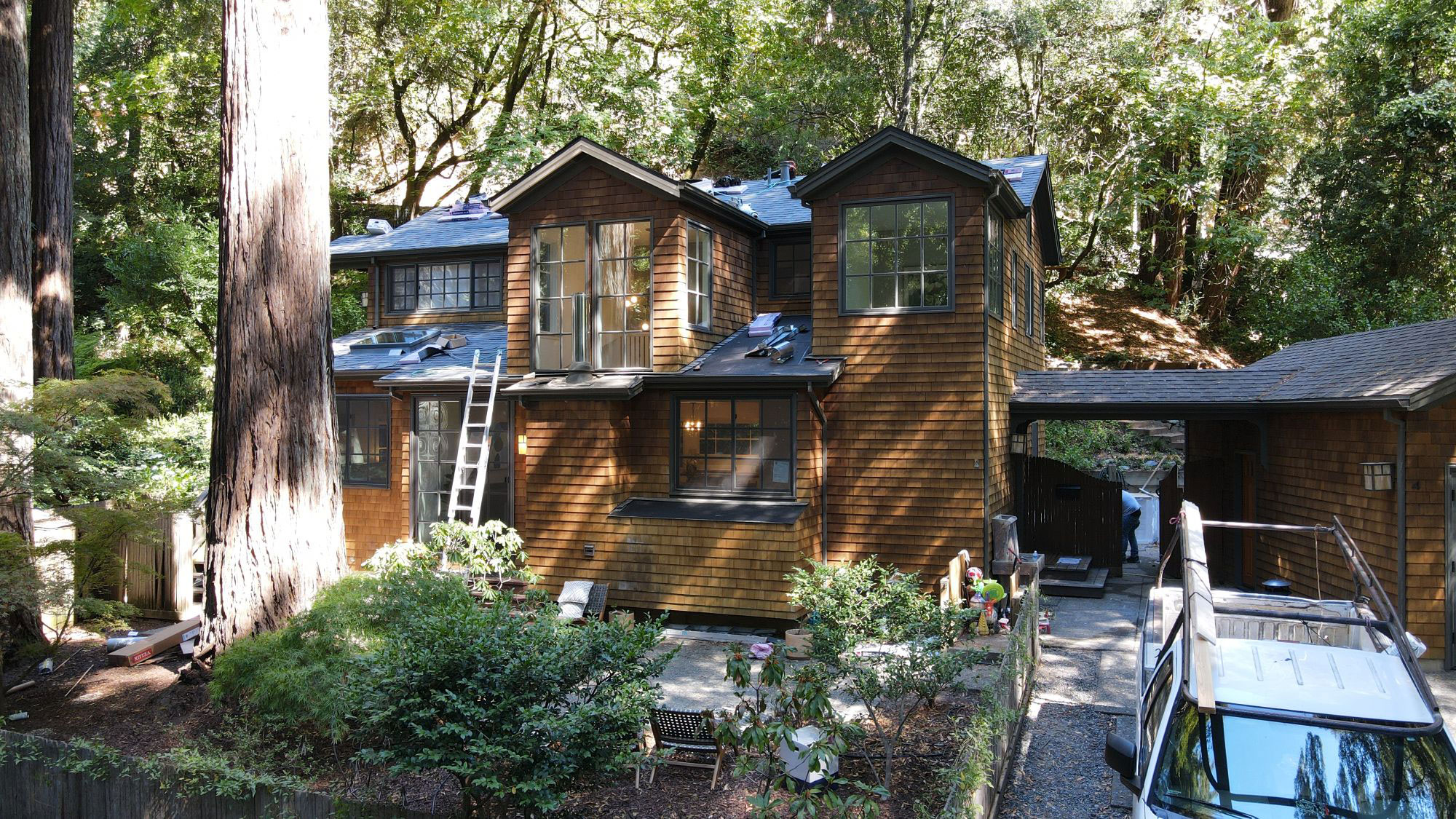 The image shows a two-story residential house with a dark roof and multiple windows, situated on a street lined with trees.