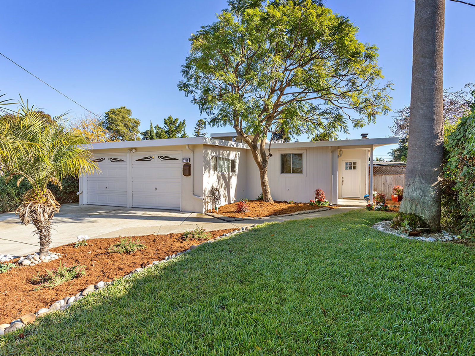 This image depicts a single-story house with a garage door, surrounded by landscaping, including a lawn and various plants, on a sunny day with clear skies.