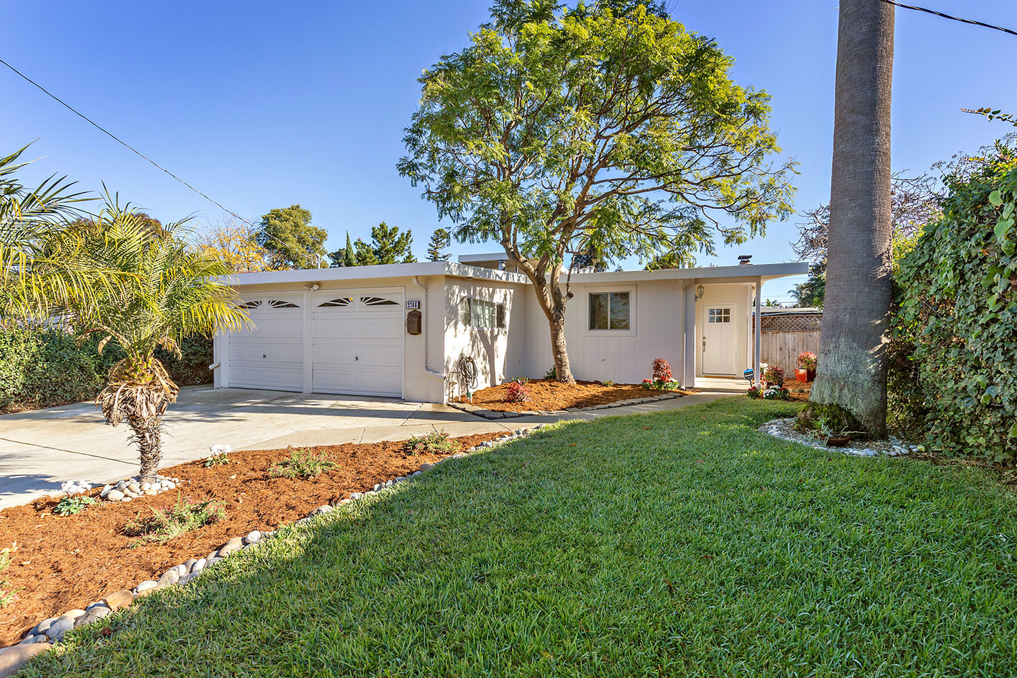 A single-story house with a white exterior and a two-car garage, set on a landscaped lawn with palm trees and a clear blue sky above.