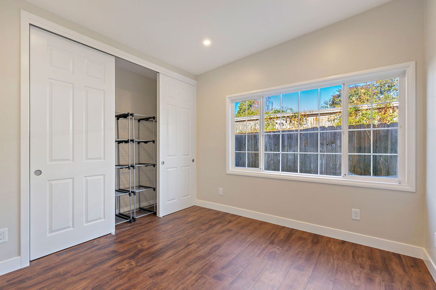 The image shows an interior space with a closed door on the right side, leading into another room. The room has a wooden floor, a window with blinds partially drawn, and a view of the outside through the window. There s a white wall visible, and a rack or shelf unit is mounted on the wall to the left of the window.