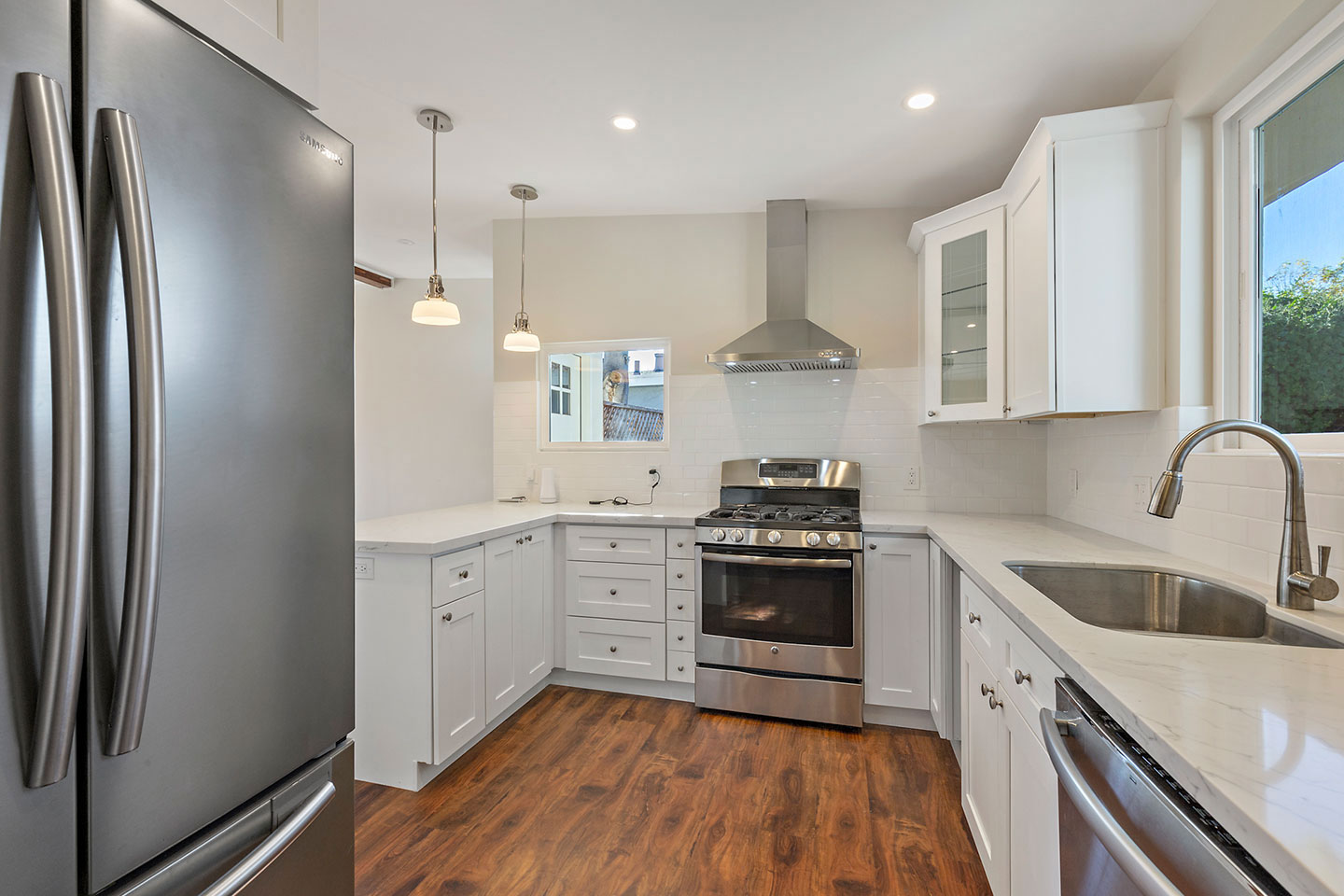 The image shows a modern kitchen with stainless steel appliances, including a refrigerator, oven, and sink, white cabinets, a hardwood floor, and a silver faucet.