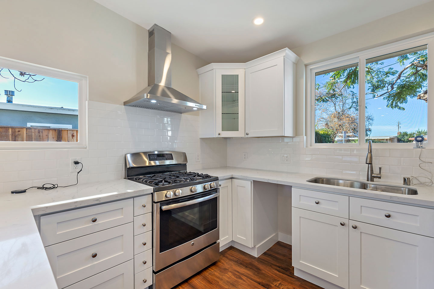 The image shows a modern kitchen with white cabinets, stainless steel appliances including an oven and refrigerator, and a large island with a sink at its center. The kitchen has a tile backsplash and a window that offers a view of the outdoors.