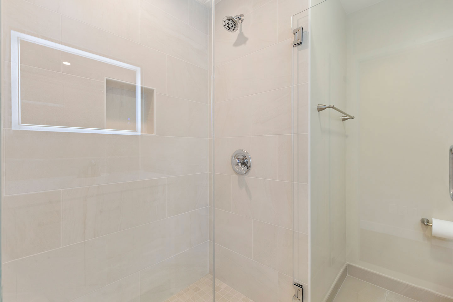 This image depicts a modern bathroom interior with a walk-in shower stall featuring clear glass doors, white tiled walls, and floor, along with a silver shower head and handle, a white toilet paper holder, and a recessed niche for toiletries.