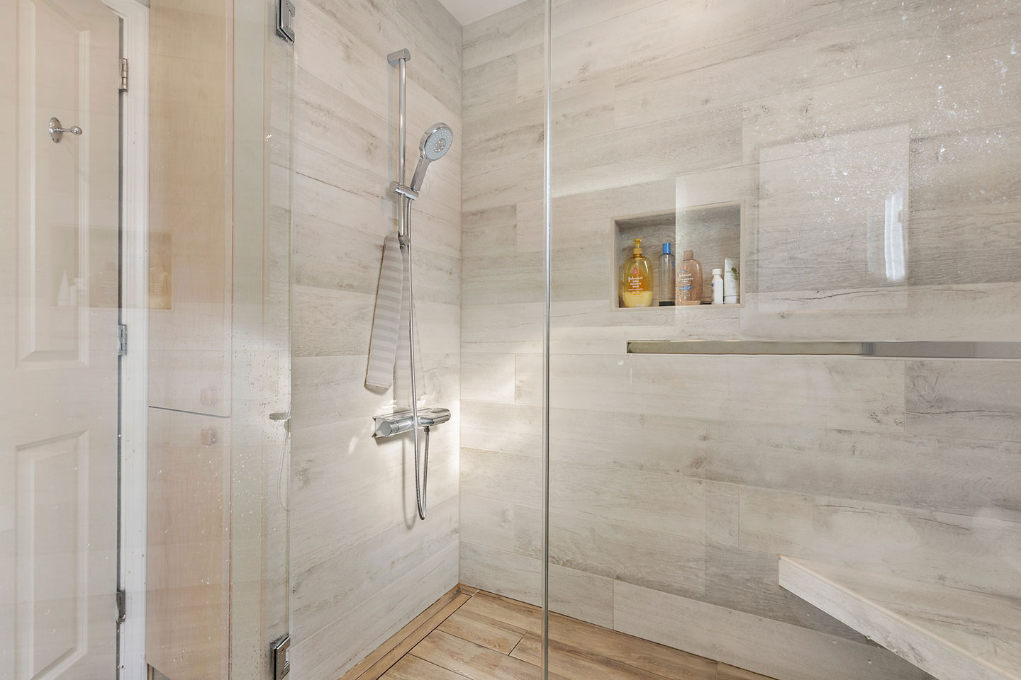 The image shows a modern bathroom with a walk-in shower featuring a glass door, a gray tiled floor, and a silver shower head.