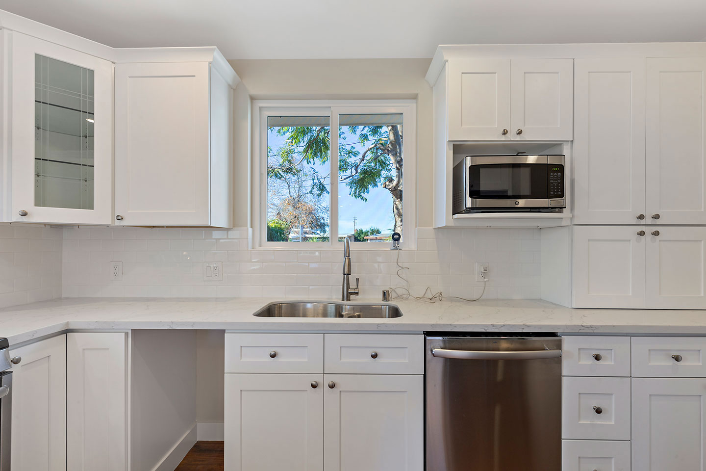 The image shows a modern kitchen with white cabinets, stainless steel appliances including a refrigerator, microwave, oven, and dishwasher, a sink, a window with a view of trees, and a countertop.