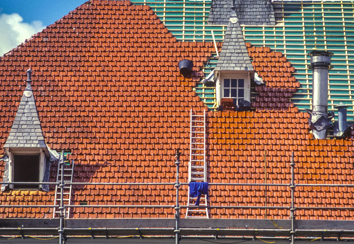 The image shows a building under construction with a damaged roof, featuring red tiles and scaffolding on the right side.