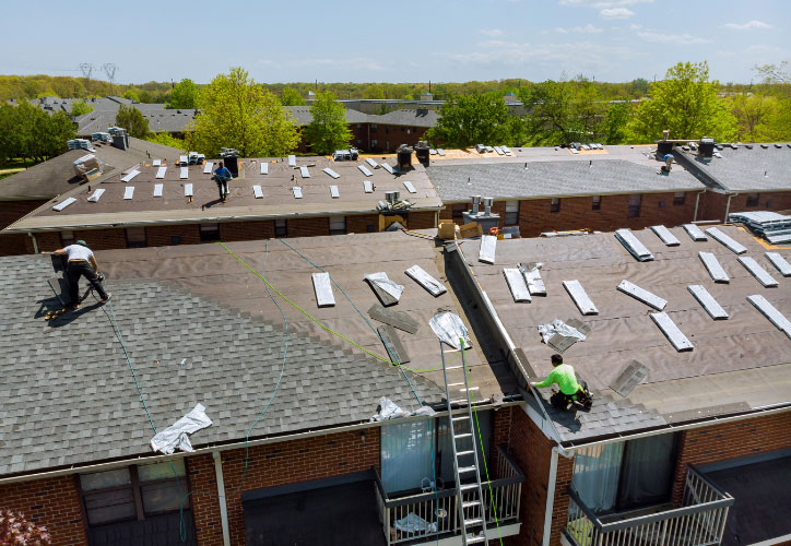 The image shows a roofing project underway on a residential building, with workers installing solar panels on the partially completed structure.