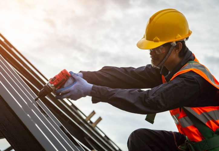 The image shows a construction worker wearing a hard hat and safety gear, working on installing solar panels on a building roof under bright sunlight.